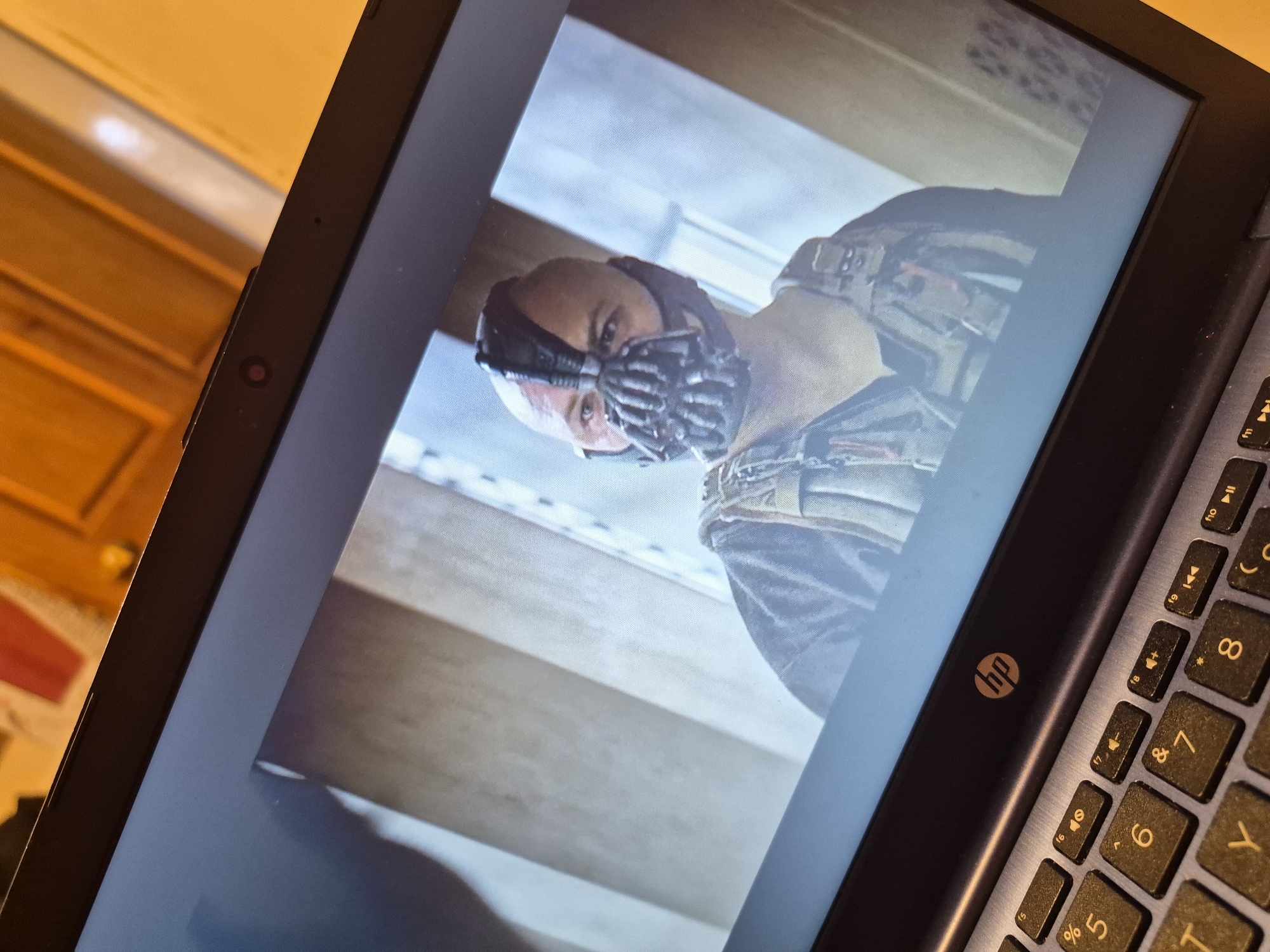 The image is a close-up, high-angle shot of a laptop screen displaying a scene from the movie "The Dark Knight Rises." A character, identified as Bane, is prominently featured, wearing his signature mask. The laptop's keyboard is visible in the foreground. The background of the image on the laptop screen appears to be an indoor setting with muted lighting. The overall scene is a still from a film being viewed on a personal device. The location within Sandwell, United Kingdom, is not discernible from the visual information provided in the image.