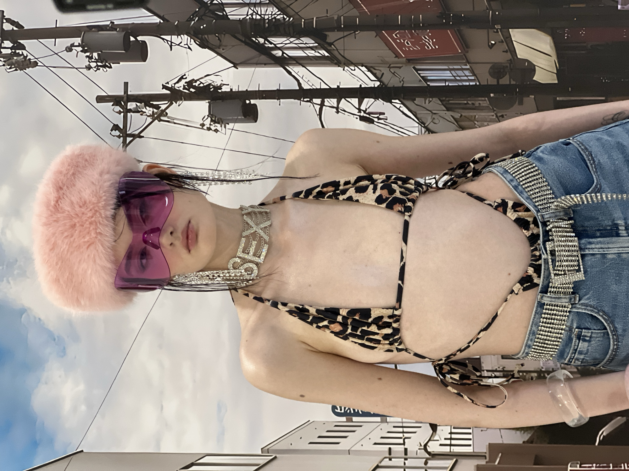 The image features a person, likely a young woman, posing outdoors in an urban setting. The subject is oriented horizontally across the frame, with her head towards the left. She is wearing a distinctive ensemble: a fluffy pink hat or headband, large purple-tinted shield sunglasses, and a leopard-print string bikini top with tie closures. A prominent, sparkly silver-colored choker necklace spells out "SEX" around her neck. She also wears blue denim jeans or shorts, which appear to be low-rise, cinched with a wide, rhinestone-studded belt. A thick, clear bangle adorns her right wrist.
The background shows a cluttered urban environment with numerous utility poles and a dense network of power lines against a partly cloudy sky. Buildings with some indistinct architectural details and signage are visible behind the subject. The lighting indicates it is daytime. The overall scene is consistent with a bustling city street, aligning with the provided location context of Khimki, Russia. The person is looking directly towards the camera, adopting a posed stance. The most prominent visible text is "SEX" on the choker necklace; other text on background buildings is not clearly legible.
