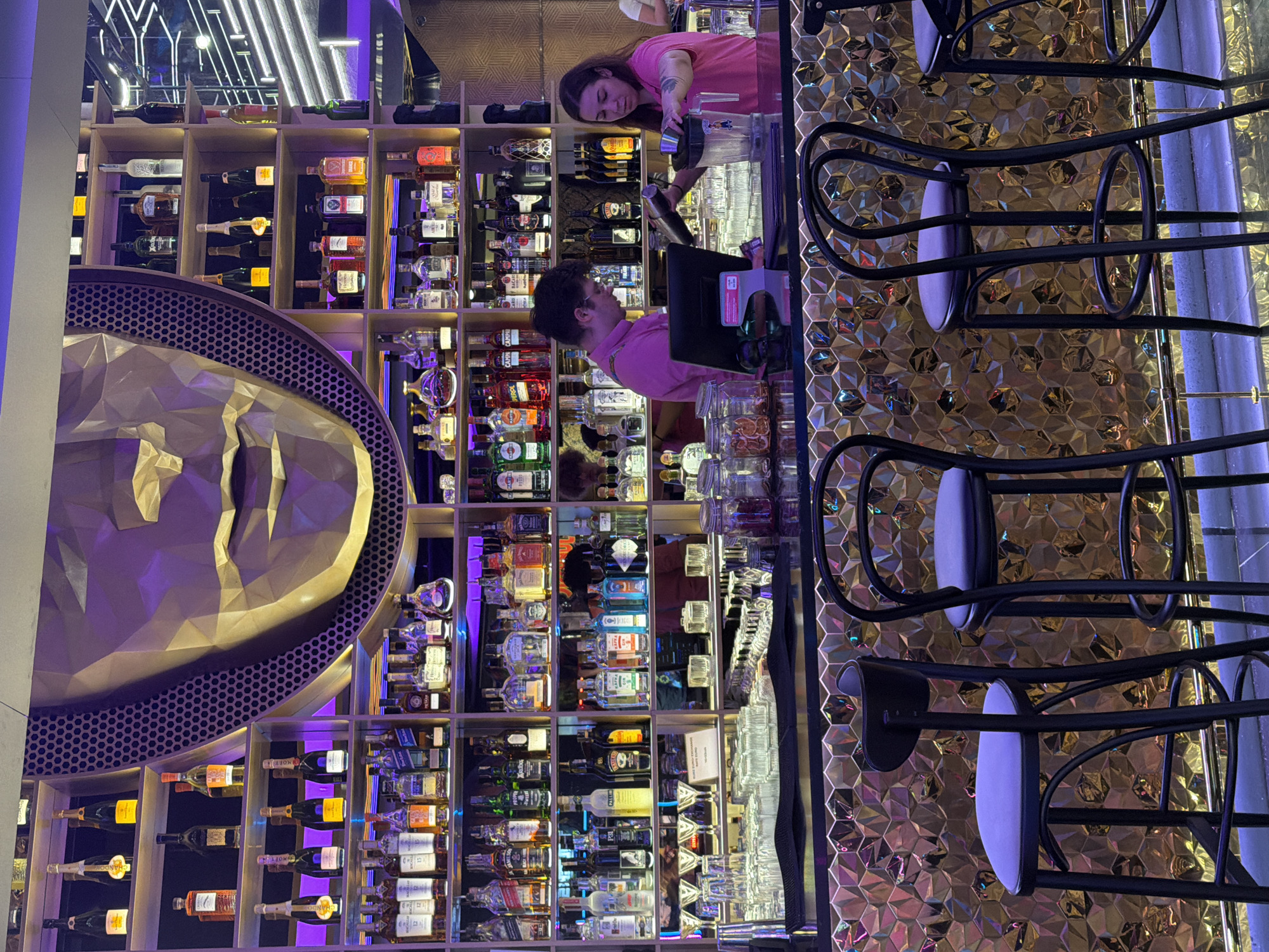 The image depicts a bar interior with a prominent golden, geometric face sculpture behind the bar. The bar is stocked with numerous bottles of liquor and beverages, arranged on illuminated shelves. Two bartenders, a man in a pink shirt and a woman also in a pink shirt, are visible working behind the counter. The man is looking at a laptop screen, while the woman is preparing a drink, holding a shaker and a jigger. Bar stools are placed in front of the counter, suggesting a seating area for patrons. The overall ambiance appears to be modern and upscale, with purple and gold lighting adding to the mood. The provided information indicates this location is in Prague, Czechia.