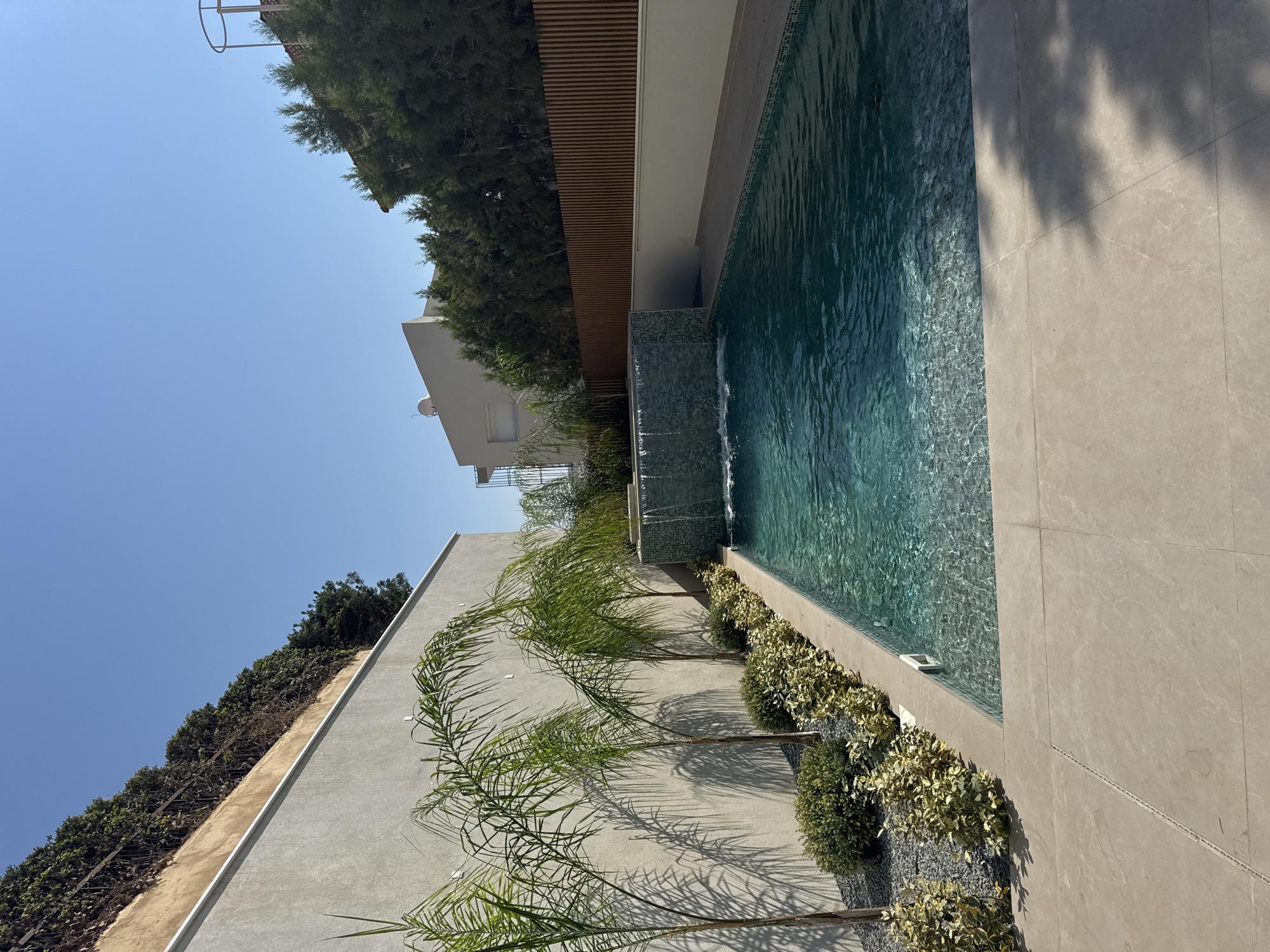 An outdoor swimming pool area is depicted under a clear blue sky in Germasogeia Municipality, Cyprus. The pool features turquoise water with visible ripples and a tiled edge. A waterfall cascades from a mosaic-tiled wall at the shallow end of the pool. To the left of the pool, a light grey wall forms the backdrop, with a row of palm trees and low-lying green and variegated shrubs planted along its base. The ground around the pool is paved with large, light-colored rectangular tiles. In the background, a wooden slatted fence runs along a raised planter filled with dark green evergreen trees. Beyond the fence, a modern white building with a satellite dish is visible, as are more trees and sections of other buildings and structures. The overall scene is one of a well-maintained private outdoor recreational space.