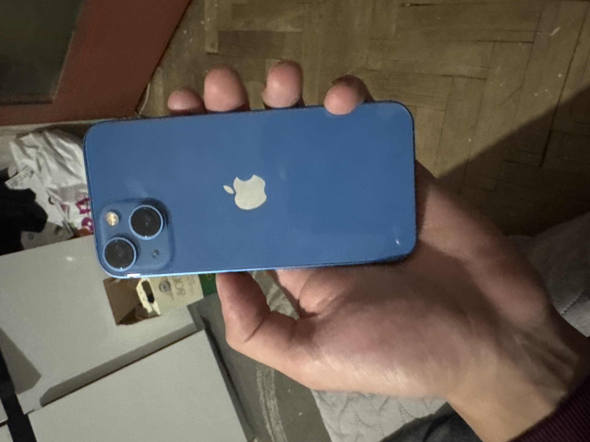 A person holds a blue Apple iPhone 13 in their hand. The phone's back is visible, showing the Apple logo and dual-camera system. The background includes a cluttered area with what appears to be a white cabinet, possibly near a window with a red frame. The setting also shows a section of a wooden floor and a piece of fabric. The location cannot be determined to be in Sofia, Bulgaria, based on the visual data provided.