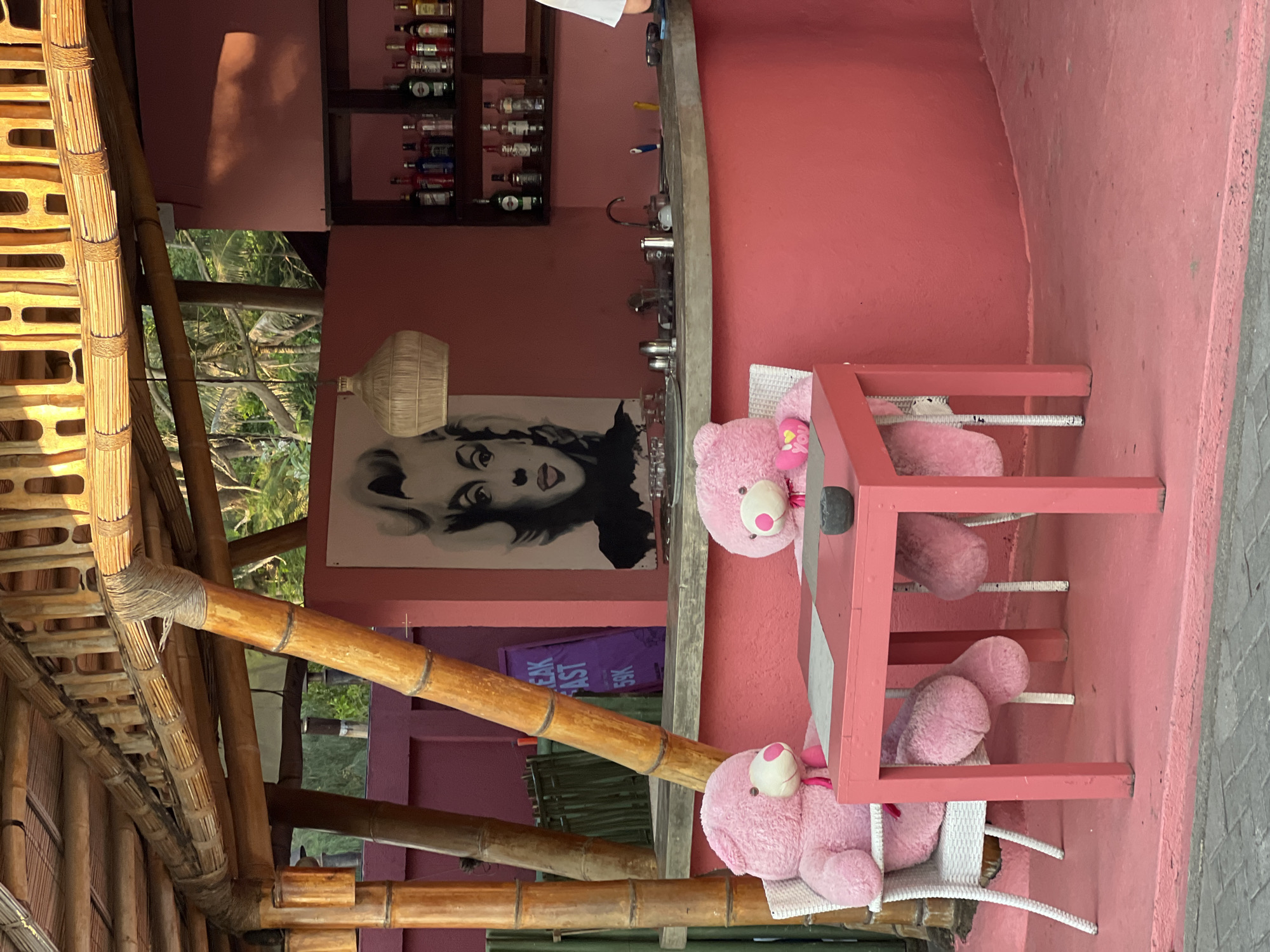 The scene is a bright pink restaurant or café with a quirky, tropical vibe. Two pink teddy bears sit at a pink table, each in a white chair. One bear has a "You" heart. The building's structure features bamboo elements, creating a rustic atmosphere. Inside, there is a bar with shelves stocked with bottles, and a black and white portrait of Marilyn Monroe on the wall. A sign that says "Break Fast 59K" is visible. The exact location is unknown, but the environment suggests it is somewhere in Indonesia.
