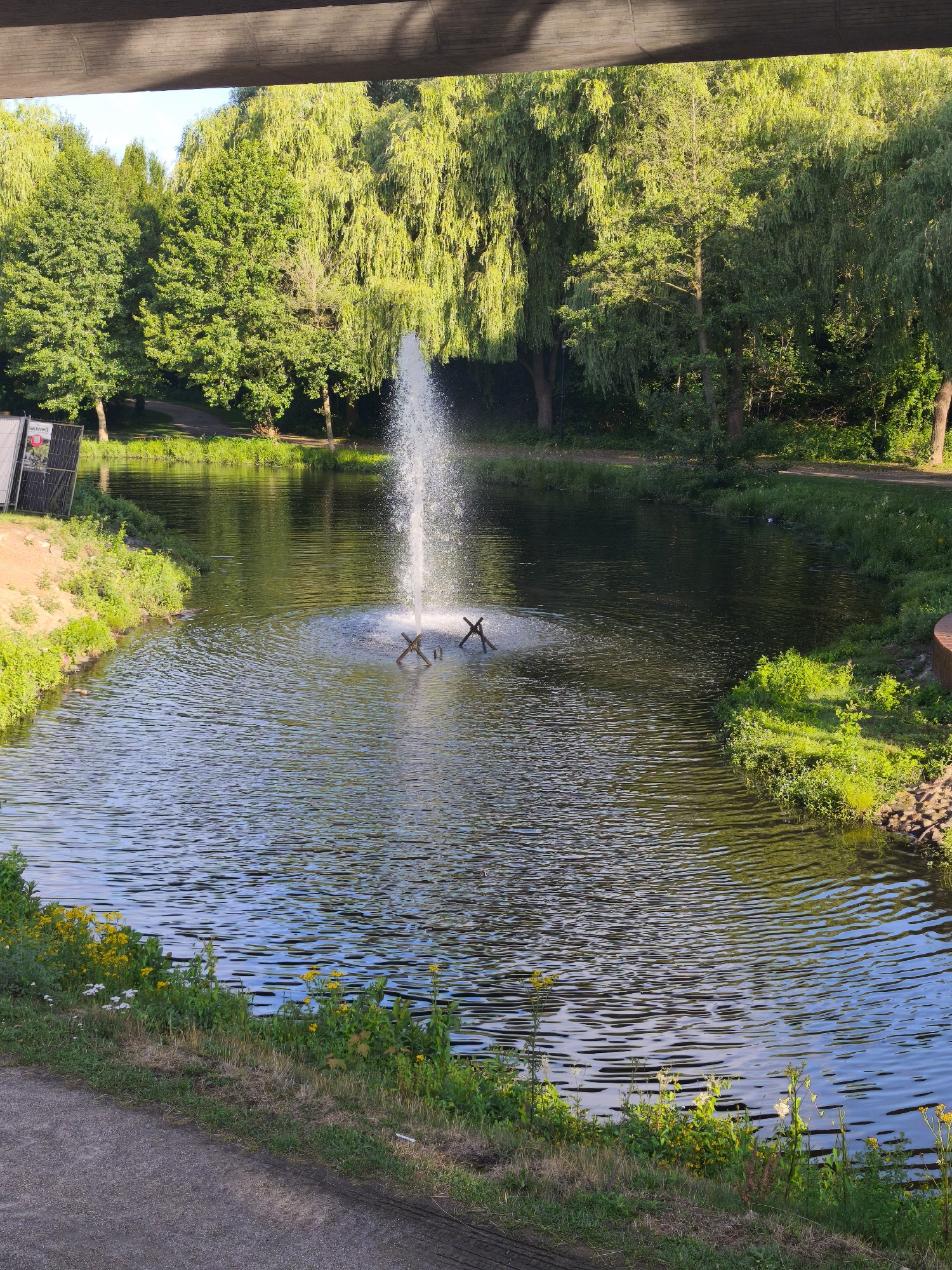 The image depicts a tranquil canal in Schwarzenbek, Germany, lined with lush green trees and vibrant vegetation. A fountain stands in the center of the canal, its water gracefully spraying upwards, creating a mesmerizing ripple effect on the water's surface. The surrounding area is a picturesque park, with a paved walkway running alongside the canal, inviting visitors to stroll and enjoy the natural beauty. The presence of a few scattered yellow flowers adds a touch of color to the scene, enhancing its serene ambiance. The overall impression is one of peace and serenity, a perfect escape from the hustle and bustle of city life.