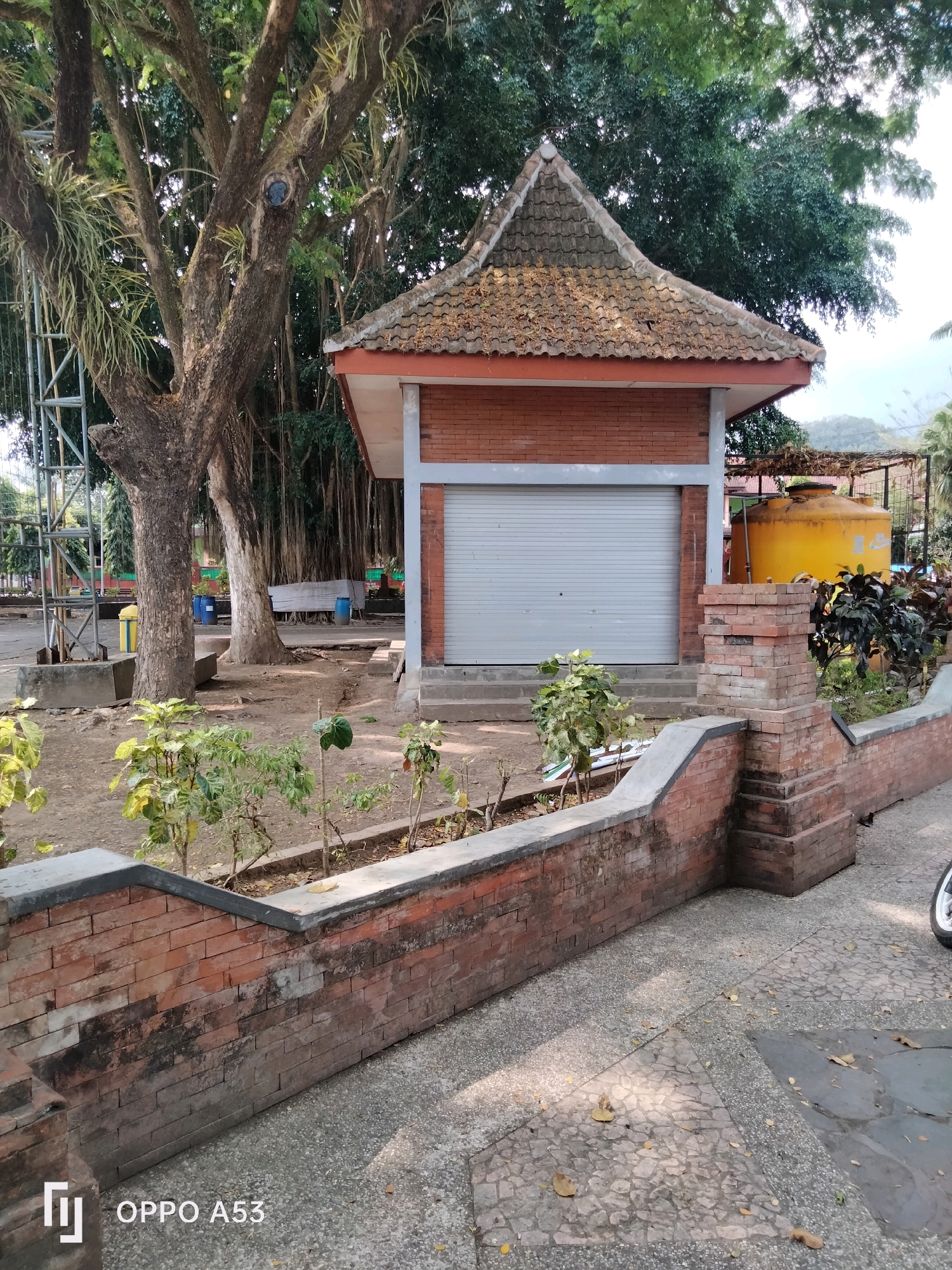 A brick building with a grey metal roll-up door and a tiled roof stands in an outdoor area. The building is surrounded by a low brick wall with a concrete top. Several trees, including a large banyan tree with aerial roots, are visible. A yellow water tank is situated to the right of the building. The ground is paved with a textured material and scattered with fallen leaves. The scene appears to be in a park-like setting in Trenggalek, Indonesia. The image has the "OPPO A53" watermark in the lower left corner.