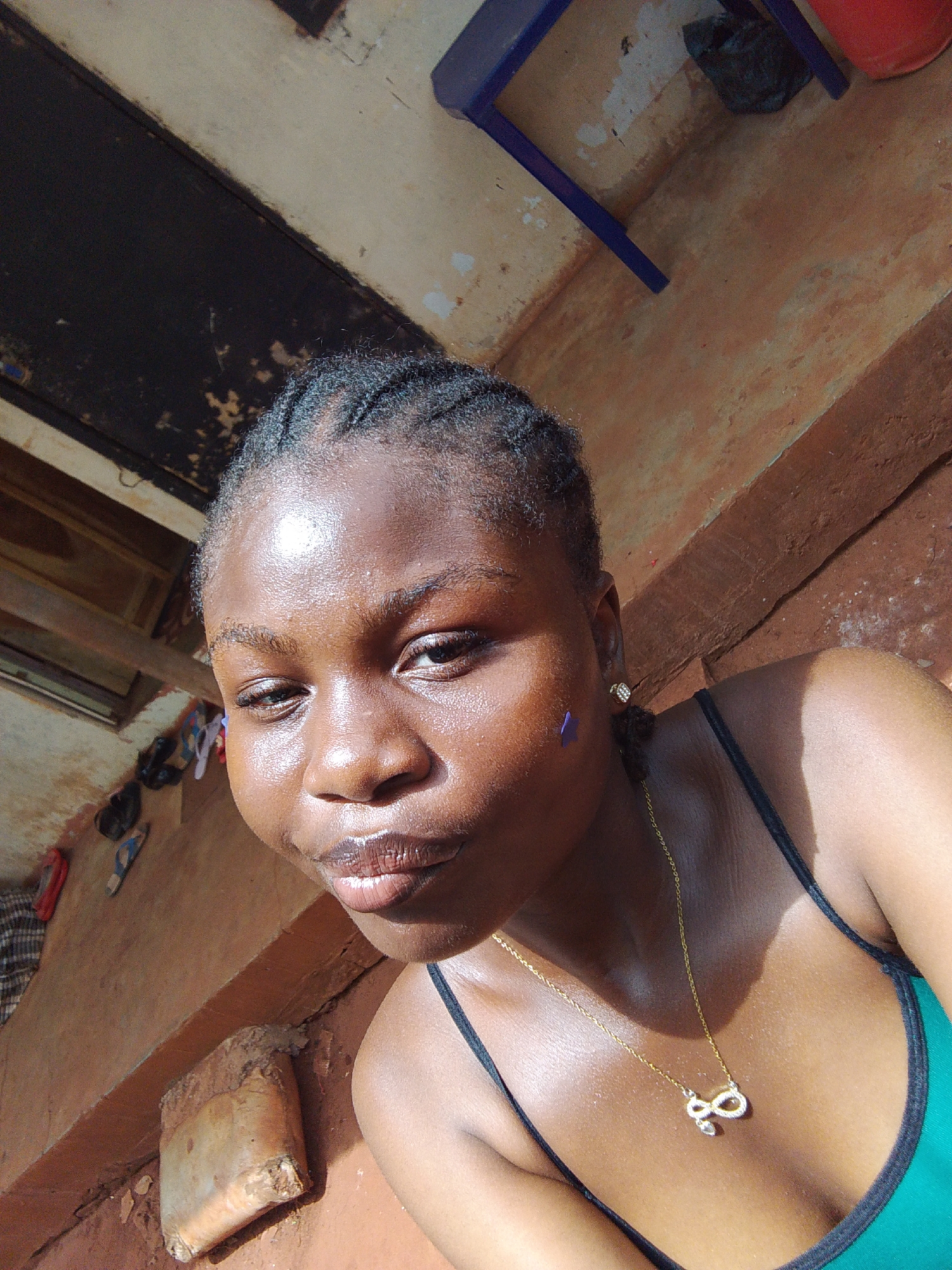 A young woman with dark skin and braided hair is positioned in a selfie shot. She wears a green tank top with black straps and a gold necklace with a pendant. A small blue mark is visible on her cheek. The background shows an interior space with walls painted in tan and brown hues. An unidentifiable black object is hanging on the wall. There are other items scattered in the background: sandals and a blanket and a small blue chair. The location is suggested to be in Nsukka, Nigeria.