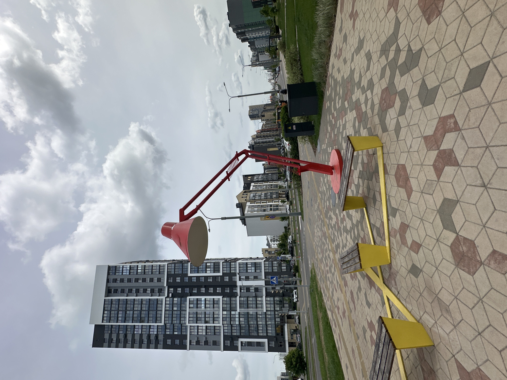 The image shows a public space in Бараўлянскі сельскі Савет, Belarus, featuring a large, red, articulated lamp sculpture next to a yellow, angular bench on a patterned, geometric-tile walkway. In the background, there is a modern high-rise building with a grid-like facade of windows and panels, as well as other buildings and a street with light traffic. The sky is overcast with clouds.