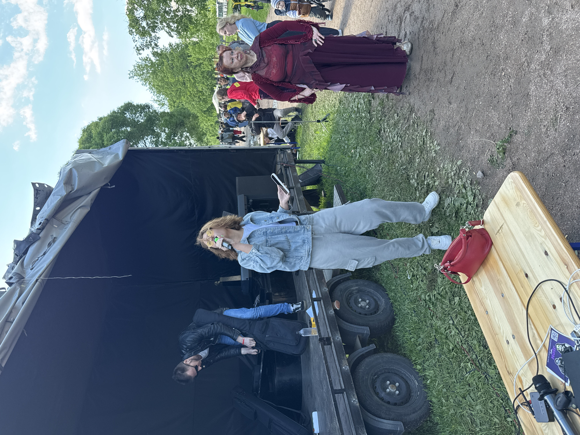The image captures an outdoor scene in Saint Petersburg, Russia, featuring several individuals and equipment indicative of a performance or event setup. A woman in a long maroon dress stands on a dirt path, facing forward and looking slightly to her right. Adjacent to her and at a lower elevation is a group of people, possibly an audience. In the foreground, a partially covered flatbed trailer serves as a makeshift stage. A woman in light-colored clothing and yellow-tinted glasses stands on the trailer, holding a microphone and looking at a smartphone. Beside her, a man in a black leather jacket and sunglasses leans over musical equipment. A wooden table with electronic equipment sits to the right of the trailer, with a red handbag on top. Cables and wires are visibly connected to the equipment on the table. In the background, trees and a light blue sky with scattered clouds are visible. The overall impression is that of an outdoor event or performance in progress, with elements of both preparation and activity evident.