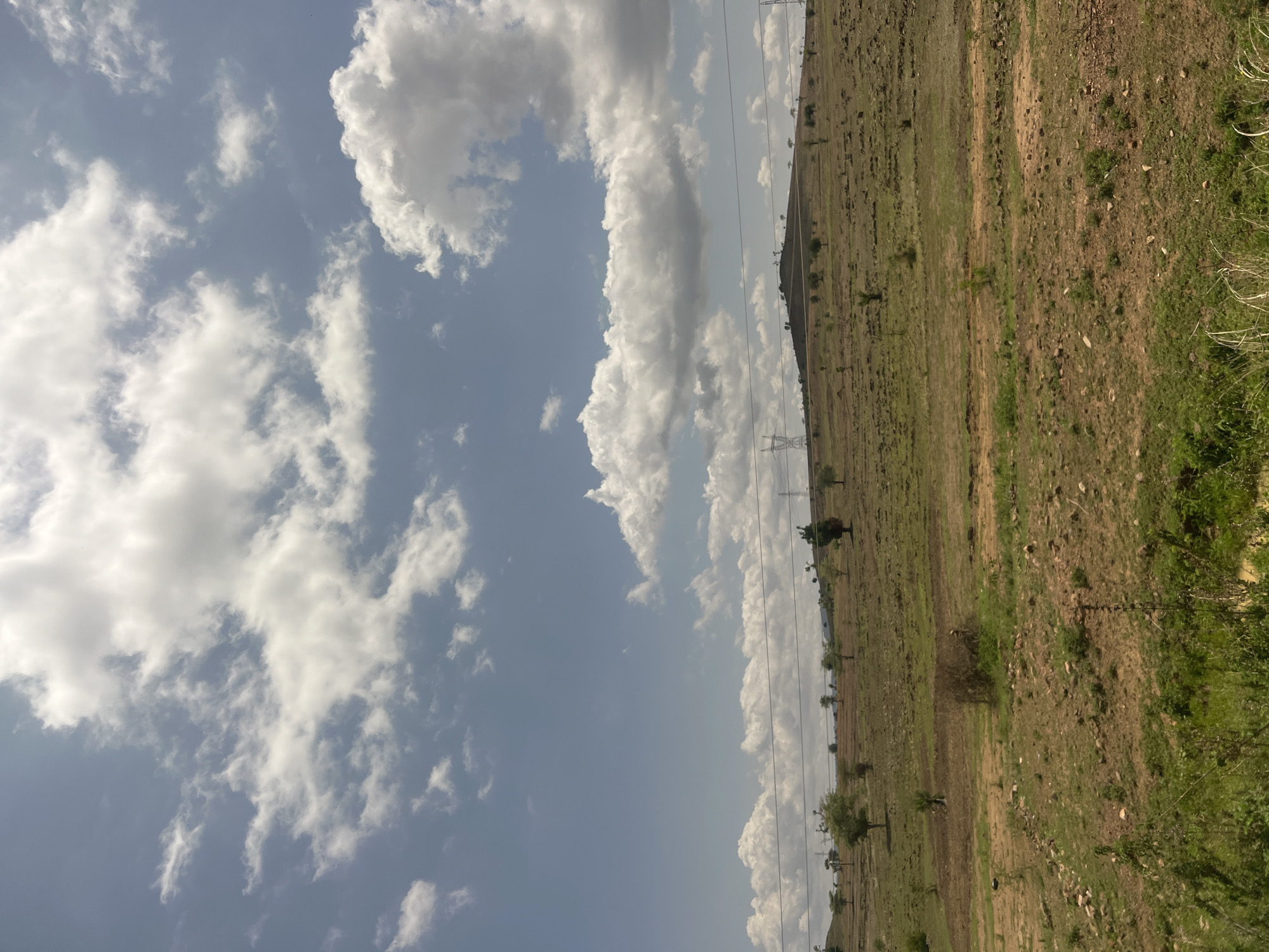 This image is a landscape photograph captured in Sabon Layi Jessu, Nigeria. The scene depicts undulating terrain under a blue sky filled with numerous scattered white cumulus clouds, some exhibiting gray undersides. The landscape comprises dry, light brown earth interspersed with sparse green vegetation and small, scattered trees, particularly on hillsides. Multiple electricity transmission pylons and associated power lines are visible across the midground and distant hills. Structures consistent with wind turbines are discernible on a distant hilltop. The foreground features uneven ground with dry, exposed soil, scattered rocks, and patches of green and dry vegetation. A visible dirt track runs horizontally across the mid-foreground. Denser green vegetation occupies the bottom edge of the frame. No people or animals are present, and no actions are depicted.