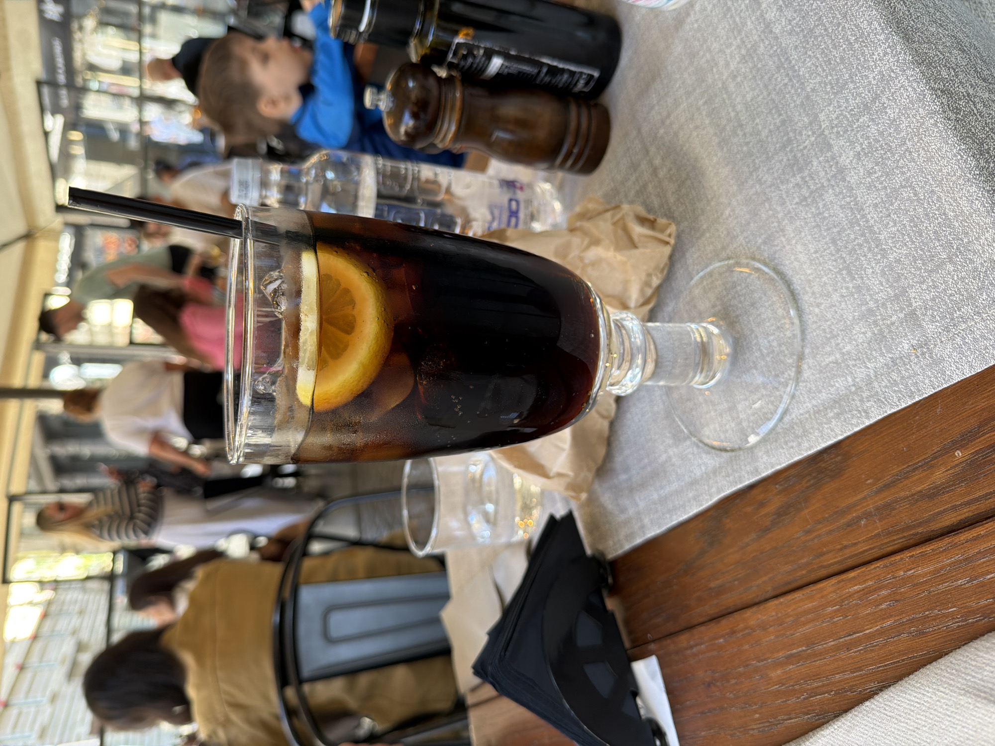 The image shows a highball glass filled with a dark beverage, presumably Coca-Cola with a slice of lemon, and a black straw, sitting on a wooden table. Adjacent to the glass are stacked black napkins. The table is partially covered with a grey cloth. Behind the glass, various objects are visible, including a clear glass, paper wrappers, and condiment bottles. In the blurred background, people are seated at tables under an awning. Based on the architectural style and overall ambiance, the location is possibly an outdoor dining area in Milan, Italy. Several patrons are visible in the background. One child is wearing a blue jacket. There is a water bottle in the background. The photograph appears to be taken at an outdoor eatery.