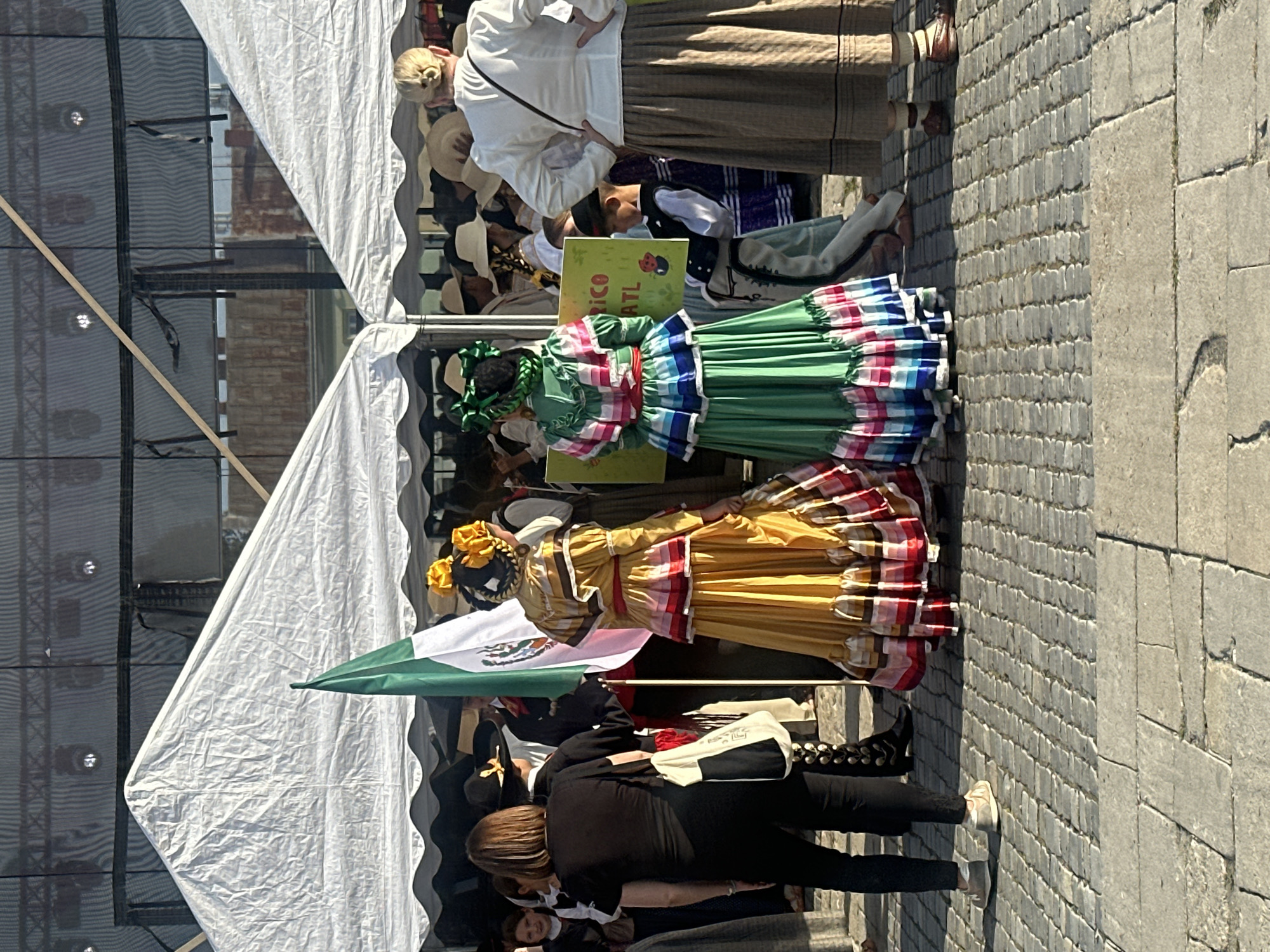 The image captures a scene at an outdoor event, likely a cultural festival, in Nowy Sącz, Poland, given the presence of a Mexican flag. In the foreground, two individuals are dressed in traditional Mexican attire. The person on the left wears a vibrant yellow dress with ruffled layers and a red sash, adorned with yellow flowers in their braided hair. The person on the right is in a green dress, also with layered ruffles and a red sash, and their hair is styled with green bows. Behind them, a Mexican flag is held aloft.
Several white tents with scalloped edges are set up, suggesting a temporary market or performance area. In the background, a darker structure with visible lighting rigs and netting appears to be a stage or backdrop. A crowd of people, some also in traditional dress or hats, are gathered, indicating an audience or participants. One person in the foreground, facing away from the camera, wears a black shirt and pants, with a white and black bag slung over their shoulder. Another individual, further back on the right, wears a white blouse and a long brown skirt, also facing away. The ground is paved with cobblestones, and a stone step is visible in the lower portion of the image. The lighting suggests it is daytime, with the sun casting shadows. A green and yellow sign with text, partially visible, is held up by someone.