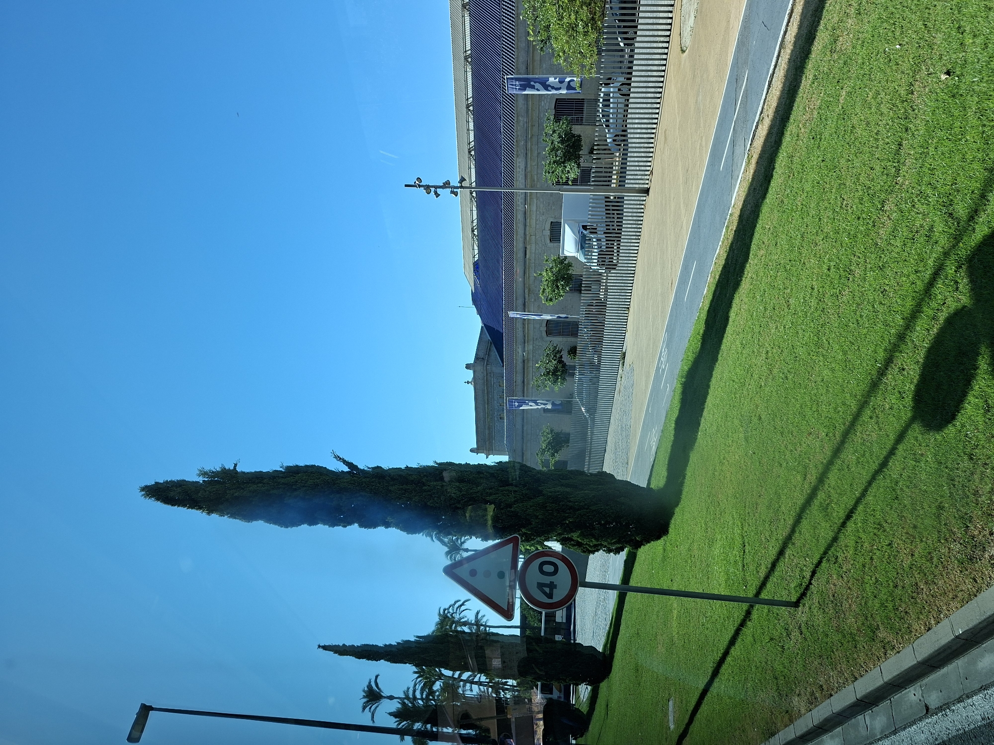 The image depicts a street scene in Alicante, Spain, under a clear blue sky. In the foreground, a patch of meticulously maintained green grass is visible, with a grey concrete curb bordering a paved area. A tall, slender lamp post stands to the left, casting a long shadow across the lawn. Two tall, narrow cypress trees are prominent features on the left side of the image. A triangular warning sign with red and white segments is positioned in front of the trees, accompanied by a circular speed limit sign displaying "40" in white on a red background. In the mid-ground, a modern building with a dark, angled roof and light-colored stone walls stretches across the frame. A metal fence with vertical bars separates the grassy area from a paved walkway and a road where a white van and a silver car are parked. Palm trees are visible in the background behind the cypress trees. The overall impression is one of a well-kept urban environment on a sunny day.