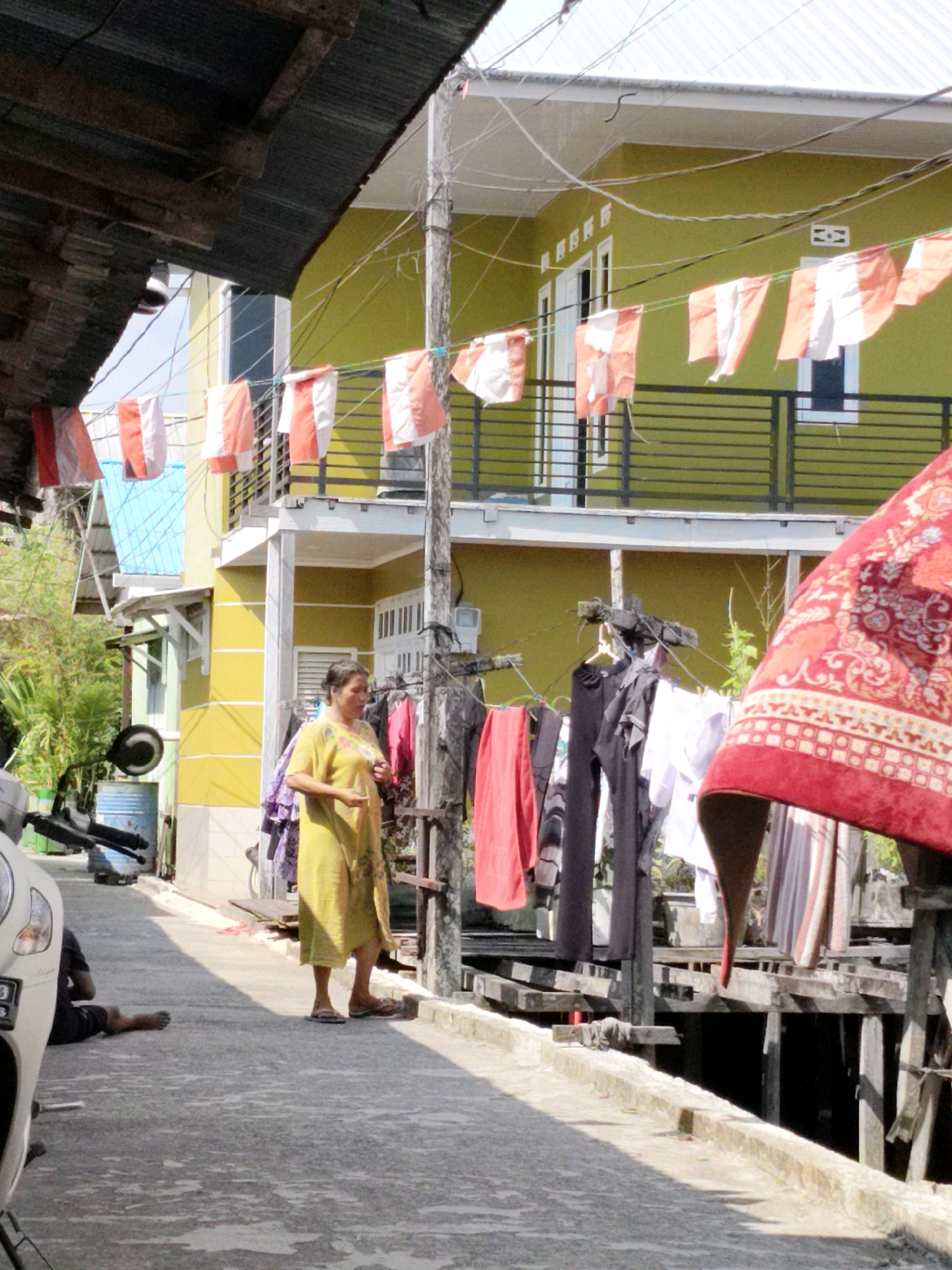 The image depicts a street scene in Balikpapan, Indonesia, characterized by a row of yellow and green buildings, some with balconies and railings. In the foreground, a woman in a yellow dress stands on a concrete walkway, seemingly engaged in an activity related to laundry, as clothes are visible hanging on a line nearby, along with a large, intricately patterned red rug draped over a railing. To the left, a portion of a white scooter is visible, and further back, a person in dark clothing is seated on the ground. The environment appears to be a residential area with a mix of traditional and modern architectural elements. The street itself is paved with concrete, and the overall impression is one of a bustling, everyday urban setting.