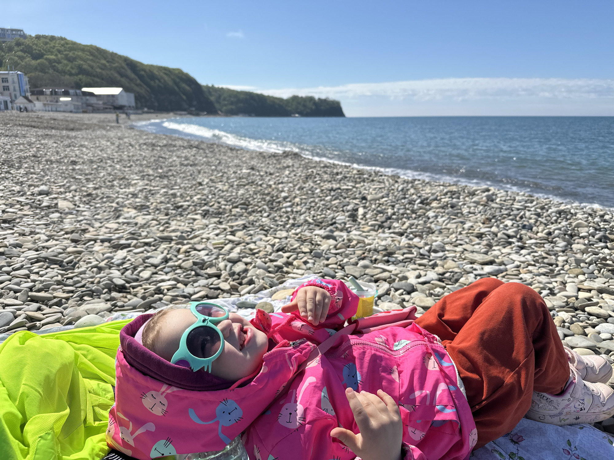 A child lies on a pebble beach in Novomikhaylovsky, Russia, wearing bright pink rabbit-print jacket, red pants, pink and white sneakers, and mint green sunglasses. They rest on a green and white patterned blanket with a lime green jacket nearby. The beach consists of small, smooth pebbles and extends to the water's edge. Beyond the water is a hilly, forested coastline, and to the left, there are buildings adjacent to the beach. The sky is blue with a few wisps of clouds on the horizon.