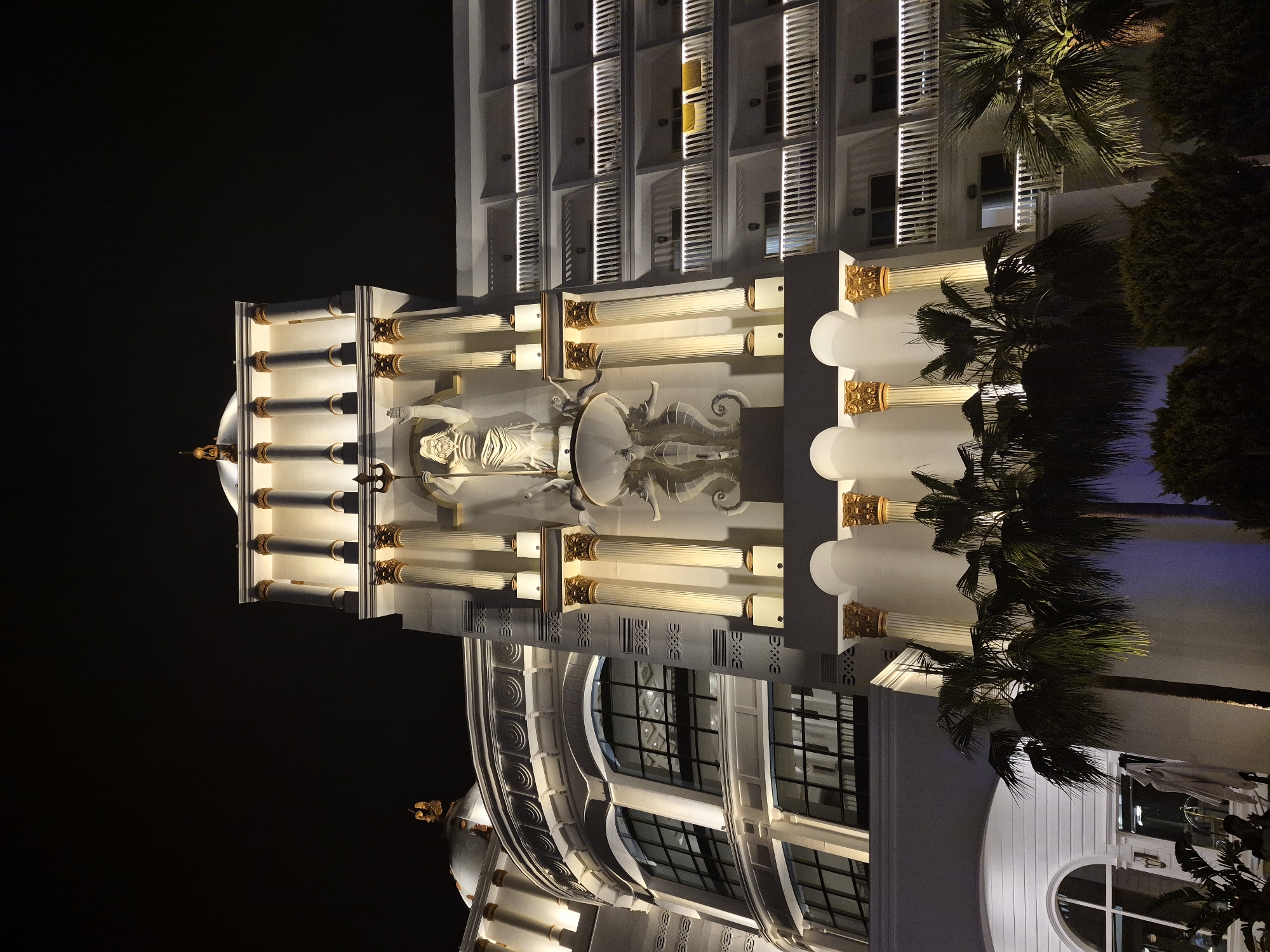 A night view of a white building with classical architectural elements in Alanya, Turkey. The building features a statue of a woman with sea creatures on a platform supported by columns. Above the statue, the building's facade has multiple stories with shuttered windows. Palm trees and other vegetation are visible on the right side of the building. The background is a dark night sky.
