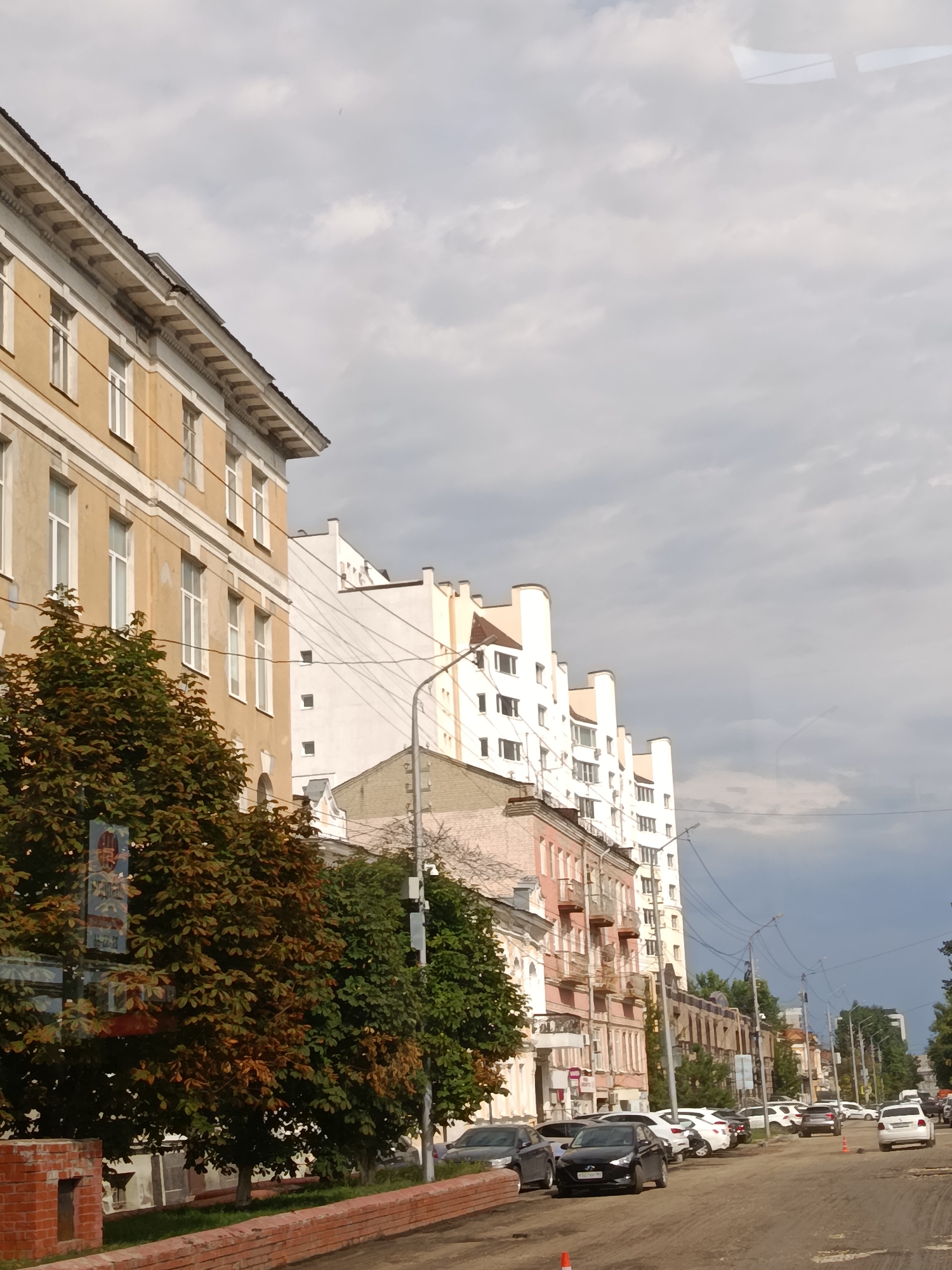 The image captures a street scene in Saratov, Russia, under an overcast sky. The perspective is from a moving vehicle, indicated by reflections on the windows and the slight tilt of the view. The scene is dominated by a row of apartment buildings, varying in architectural style and height.
On the left, a light yellow building with white trim and windows stands prominently, featuring ornate detailing along its roofline. Adjacent to it, a white building with a more modern, blocky design is visible. Further down the street, other buildings of different colors and ages line the road, some with balconies and visible windows.
The street itself appears to be under repair or construction, with a rough, uneven surface and scattered orange traffic cones. Several cars are parked along the side of the road and are also driving down the street. Among them are a black sedan in the foreground, a white sedan further down, and other vehicles further in the distance.
Trees with foliage in various stages of autumn color – greens and browns – are planted along the sidewalk, providing a natural element to the urban landscape. A brick wall and some greenery are also visible in the foreground. Overhead, power lines crisscross the sky. The overall mood of the image is functional and urban, with the cloudy weather suggesting a potentially cool day. No people are clearly visible in the frame, and no discernible text is present, except for what appears to be a blurry sign on a pole near the trees.