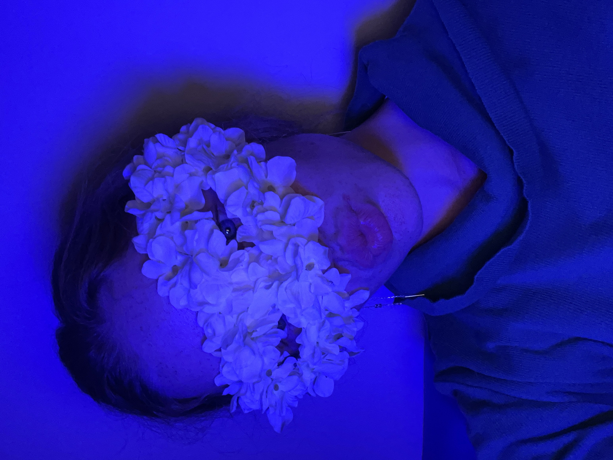 The image depicts a person lying down, with a garland of white flowers covering their eyes and forehead. Their face is illuminated by blue lighting. The individual is wearing a dark-colored garment, possibly a sweater. A metal pin is visible near their neck. The background is a solid blue color. There are no indications of the location of the city of Budva, Montenegro, or any identifying elements of a city.