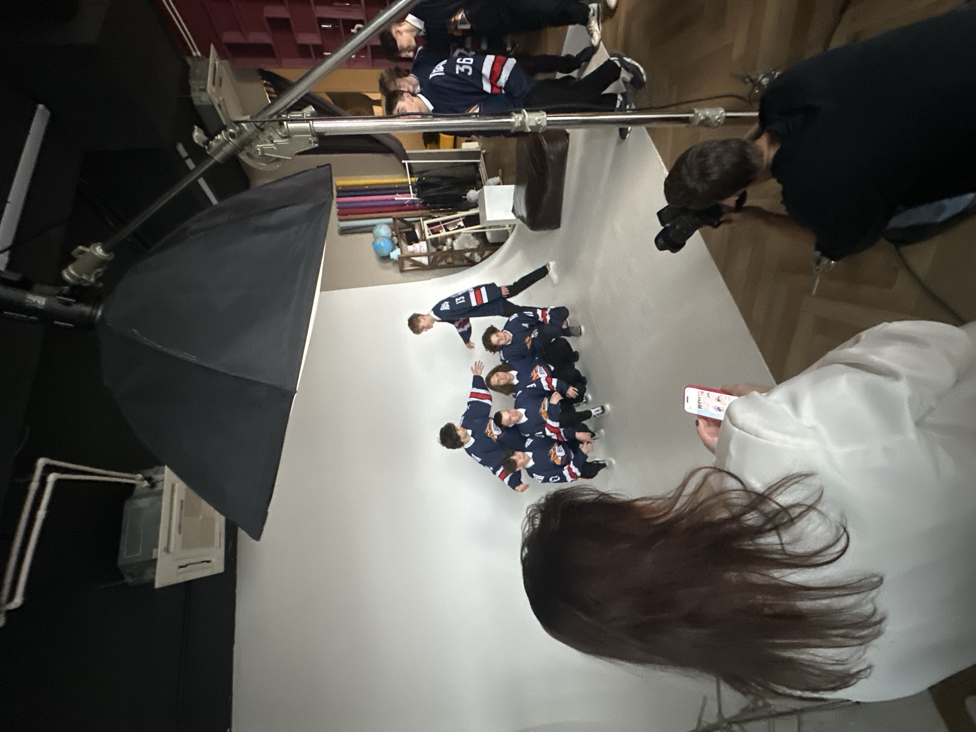 The image depicts a behind-the-scenes view of a photo shoot in what appears to be a studio. Multiple people are visible, including a group of young individuals wearing similar navy blue hockey jerseys with red and white stripes and numbers. These subjects are positioned on a white backdrop. At least one person is seen taking photos of the group, while another person, dressed in white, is holding a smartphone. A large lighting umbrella on a stand is also visible. The exact location in Unknown, Unknown, is not discernible from the image.