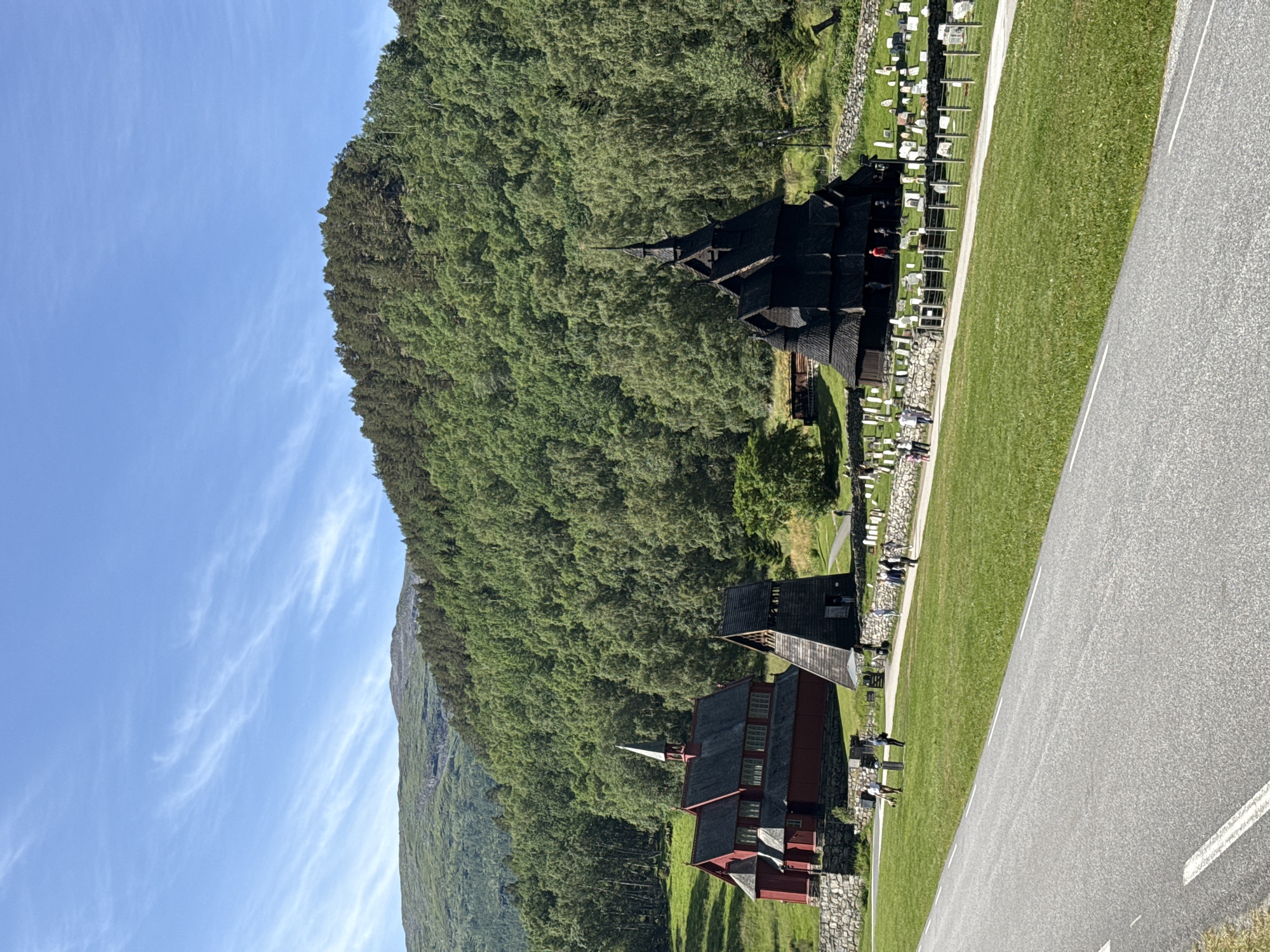 In Borgund, Norway, a paved road curves into the foreground. To the right, a green lawn is bordered by a stone wall and a white fence. Beyond the fence, a graveyard with numerous white headstones is situated. In the mid-ground, a traditional dark wooden stave church with a steeply pitched, multi-tiered roof and a tall spire stands prominently. To its left, a red wooden church with a steep roof and a bell tower is visible, also constructed with wooden walls and a stone foundation. Between the two churches, a wooden bell tower with a dark, angled roof is positioned. The background is dominated by a dense, green forest covering a mountainside, which transitions into a lighter green, grassy hill in the distance. The sky is a clear, bright blue with thin, wispy white clouds. Several people are scattered throughout the scene, walking near the churches and along the paths.
