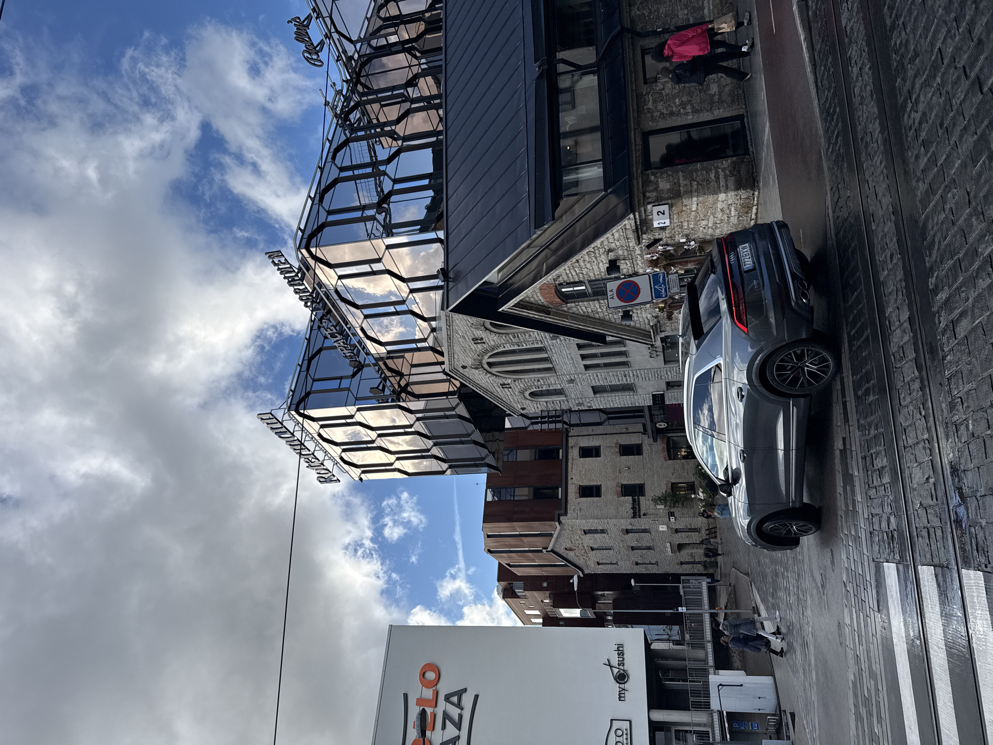 The image captures a street scene in Tallinn, Estonia, specifically in the Rotermann Quarter. A grey Audi Q8 SUV is positioned in the foreground, on a cobblestone street with tram tracks. The car is facing to the right, with its rear taillights illuminated. In the background, several buildings are visible. To the left, a modern white building with orange lettering that reads "ROTTERDAM PLAZA" stands prominently, alongside a smaller building with the text "my sushi". The central and right portions of the image are dominated by the Rotermann Quarter's distinctive architecture, characterized by a mix of old stone buildings and a modern glass-and-steel structure. The glass facade of the modern building reflects the cloudy sky. Two people are walking on the sidewalk to the right of the car. The sky above is partly cloudy, with patches of blue sky interspersed with grey clouds. The street appears wet, suggesting recent rain.