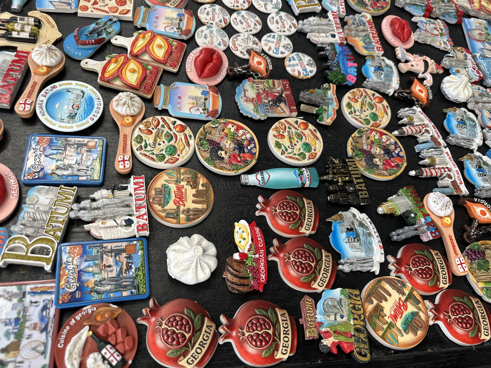 A high-angle, eye-level shot presents a multitude of souvenirs from Batumi, Georgia, arranged on a flat, dark surface. The souvenirs are of various shapes and sizes, predominantly magnets showcasing iconic landmarks, cultural symbols, and culinary elements of Georgia.
Many magnets bear the words "Georgia," "Batumi," or "Georgian Cuisine." Recurring motifs include pomegranates, Georgian dishes (like khinkali), cityscapes of Batumi, and illustrations of cultural scenes. Some magnets feature unique designs such as a pair of lips with "Kisses from Georgia," a miniature bottle of Borjomi mineral water, and replicas of buildings and monuments found in Batumi. The color palette is diverse, with prevalent shades of red, white, blue, and brown.
The composition is dense, with minimal space between the items, creating a visual array of Georgian mementos. The lighting is bright and even, ensuring all details are clearly visible. The background is out of focus, further accentuating the collection of souvenirs.