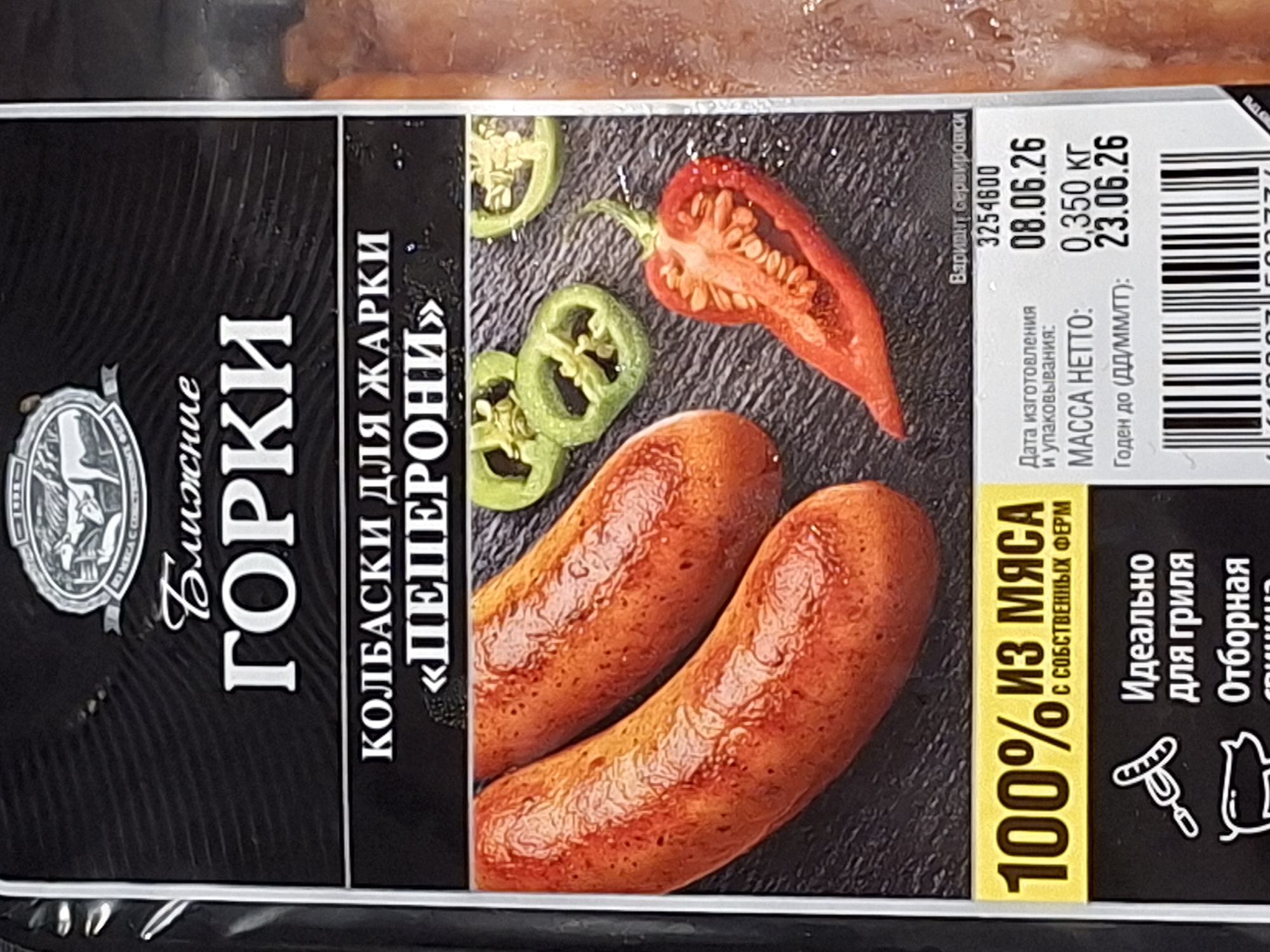 The image shows a close-up of a package of "GORKI" brand sausages, specifically "PEPERONI" flavor, made by "BLIZHNIYE" company. The packaging is predominantly black with white and yellow text and features an illustration of sausages alongside sliced jalapeños and a red chili pepper. The text indicates "100% MEAT FROM OWN FARMS" and that the sausages are "ideal for grilling." Nutritional information and production dates are also visible. The image is a product shot, likely taken in a retail or studio environment, and does not depict any specific location in Moscow, Russia, or any people.