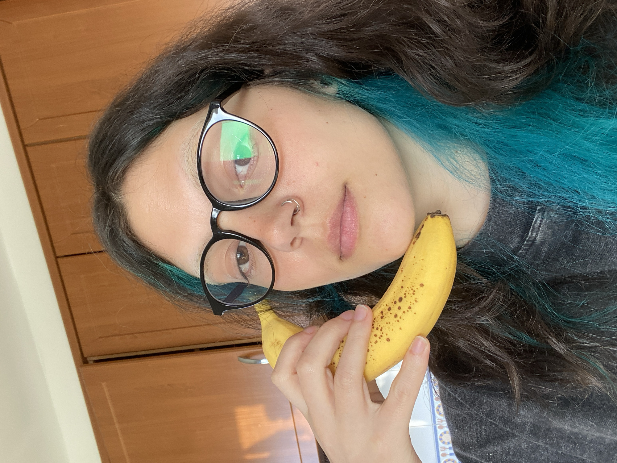 A young woman with long, dark brown hair with turquoise streaks wears round black glasses and a nose ring. She holds a ripe banana close to her face. She appears to be indoors, possibly in a kitchen, as evidenced by the wooden cabinets in the background. The location is unknown.
