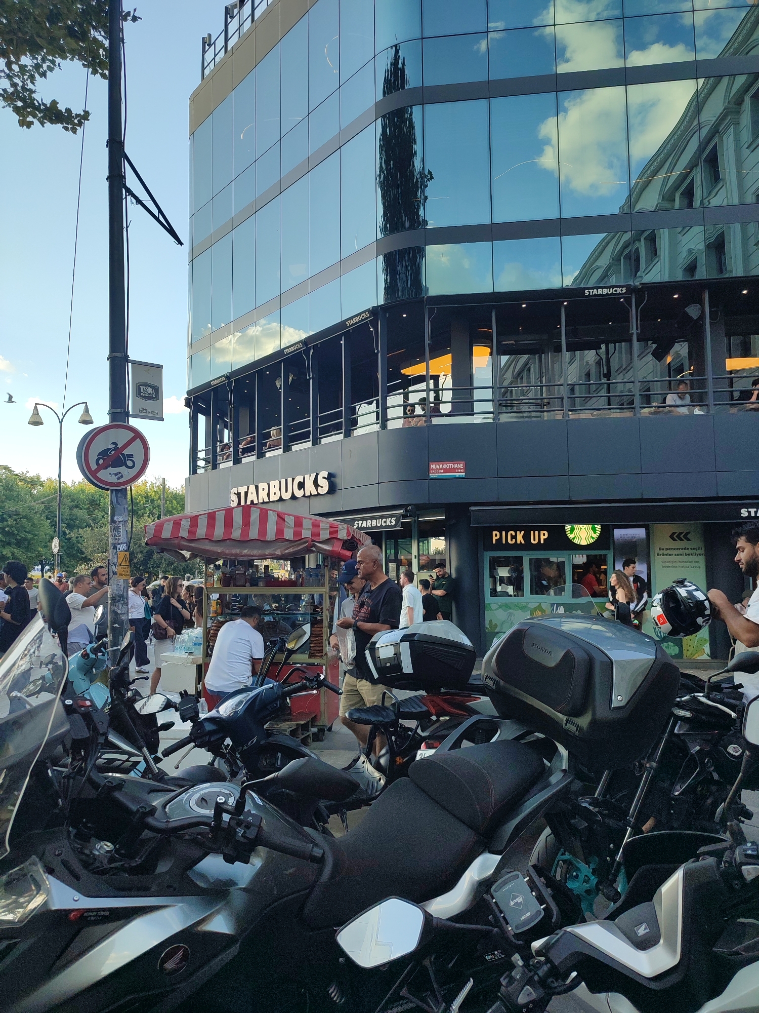 The image depicts a street scene in Kadıköy, Turkey, dominated by a modern Starbucks store with a glass facade reflecting the sky and surrounding buildings. In the foreground, several motorcycles are parked, with their handlebars and seats visible. A street vendor's cart with a striped awning is set up in front of the Starbucks, offering food items. A crowd of people is gathered around the cart and the entrance of the coffee shop, with some individuals standing, others seemingly in conversation, and one person in the foreground holding a helmet. A street sign with "Muvakkıthane" is visible on the building. A pole with a traffic sign prohibiting motorcycles is also present. The overall scene suggests a bustling urban environment on a sunny day.