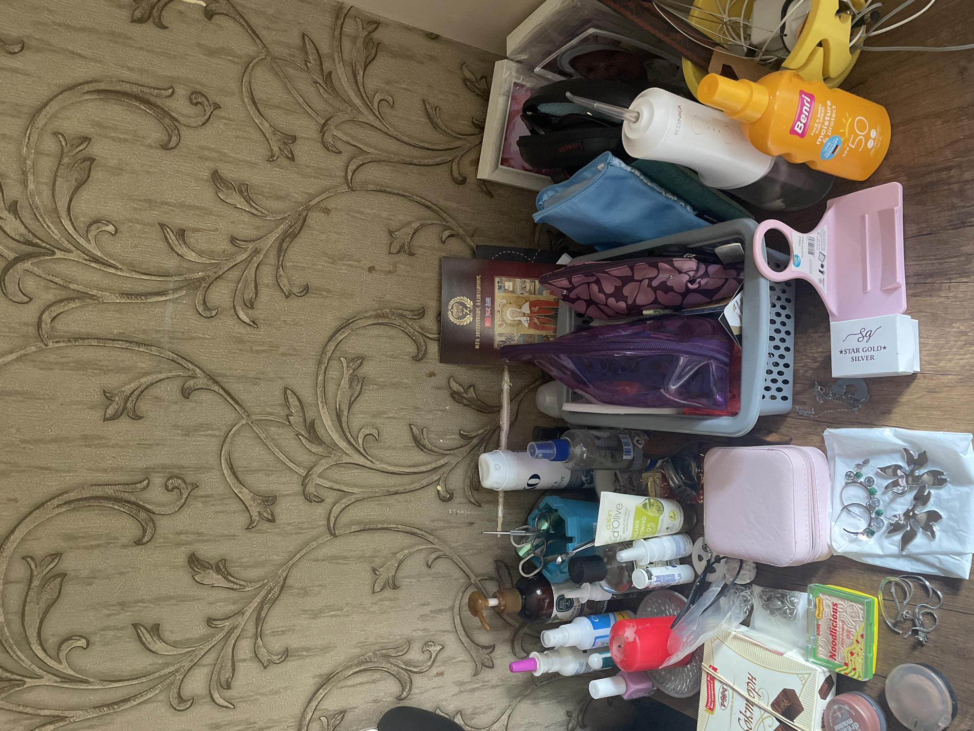 The image depicts a tabletop cluttered with various personal care items, toiletries, and accessories. The surface is a wooden table, and behind it is a wall covered with textured, patterned wallpaper featuring an ornate, floral design in shades of beige and brown.
On the table, there are several bottles and containers, including a large brown pump bottle, a white spray bottle, a blue spray bottle, and a tube of "dalan d'Olive" organic cream. A red candle is prominently displayed. There is also a pink jewelry box, a grey basket containing several items, and a variety of jewelry, including necklaces and earrings, laid out on a white paper. Among the items are makeup products, scissors in a blue holder, and a box labeled "Noodlicious." To the right of the basket, a white electronic device with the brand name "KONKA" is visible, alongside a bright orange bottle of "Benri" face and body moisture protect SPF 50 sunscreen. A pink hair clip and a white box with "STAR GOLD SILVER" text are also present.
The image appears to be taken indoors, likely in a personal space such as a bathroom counter or a dressing table. The lighting suggests it might be daytime, with ambient light from a nearby source. There are no people visible in the image. The overall impression is one of organized clutter, with a collection of everyday items arranged on a surface.
While the image captures a variety of personal items, there are no specific visual cues that definitively place this scene within Kadıköy, Turkey. The items themselves are not exclusively Turkish, and the wallpaper design is a common decorative element found globally. Without additional context or identifiable landmarks, it is not possible to ascertain the precise location within Kadıköy.