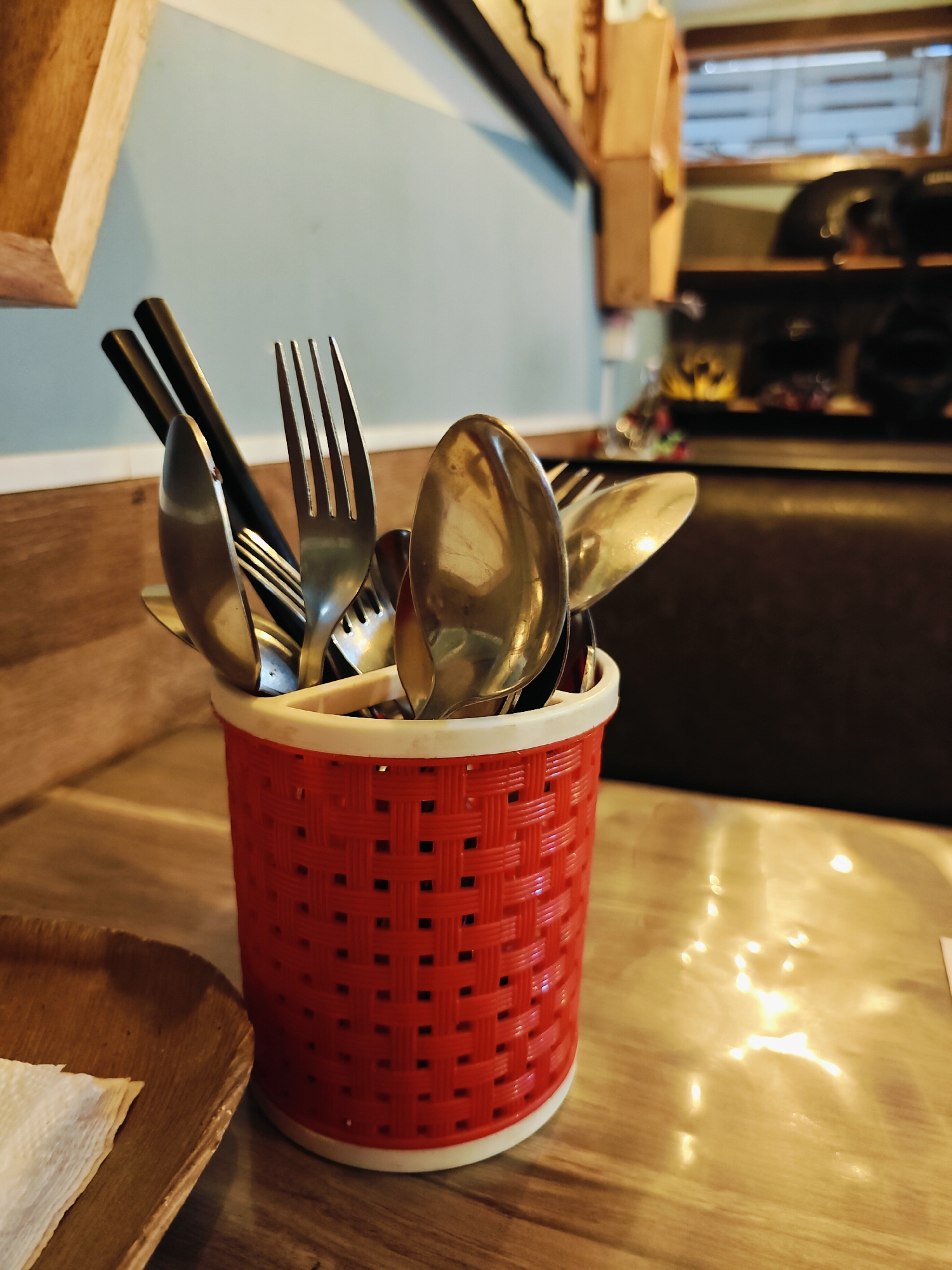 The image shows a close-up of a red woven utensil holder filled with silverware. The holder is on a wooden table, with a portion of a wooden plate and a napkin visible in the foreground on the left. In the background, there are shelves with various items, including what appear to be helmets and other restaurant-related objects. The lighting suggests an indoor setting, possibly a restaurant or cafe. The image does not provide any information about the city of Lalitpur, Nepal.