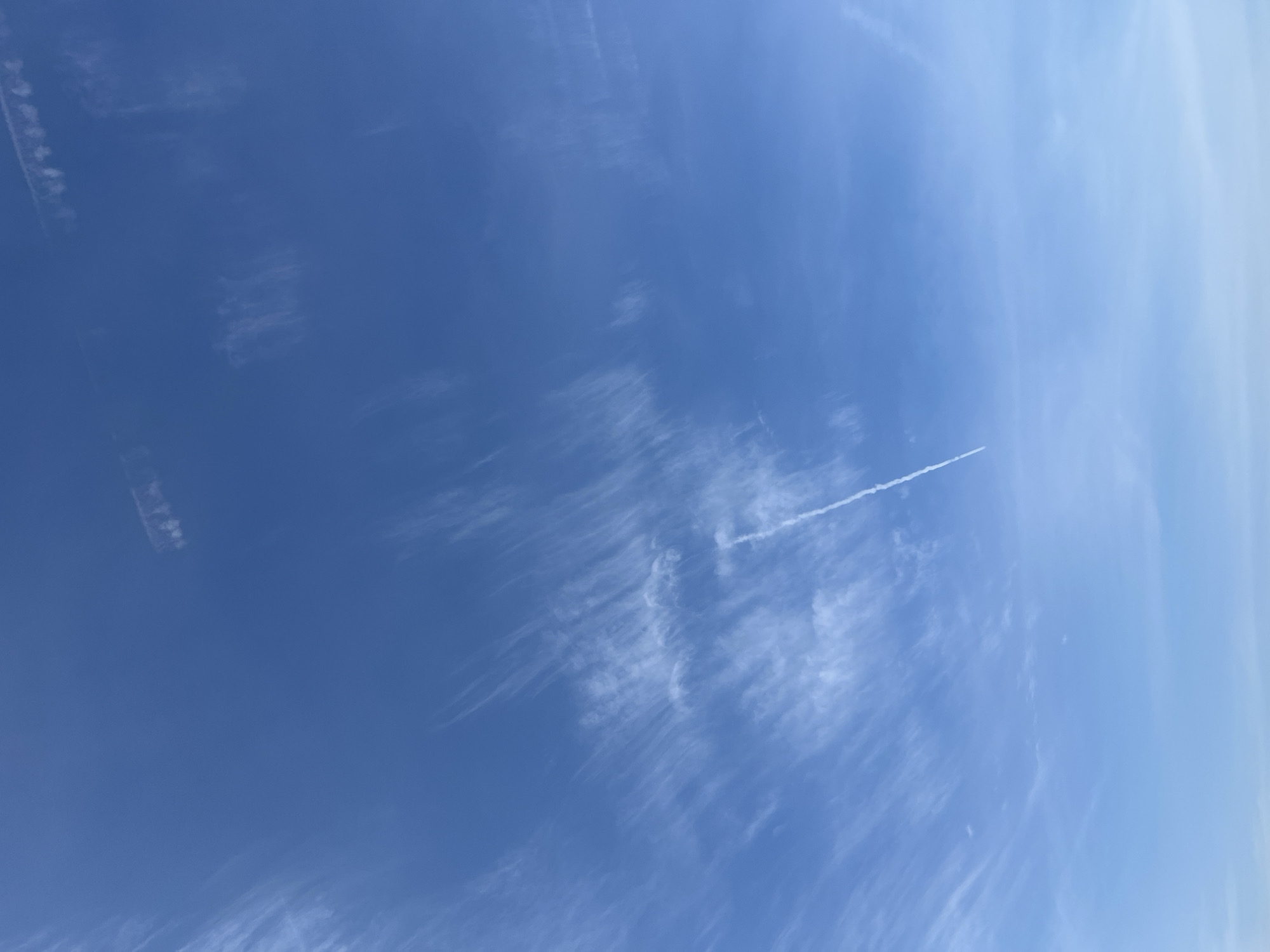 The image depicts a clear blue sky with scattered cirrus clouds. A prominent contrail, likely from an airplane, is visible stretching diagonally across the frame from the lower right to the upper left. The sky is a uniform azure, with the wispy clouds adding texture and depth. The lower portion of the image shows a slightly lighter shade of blue, suggesting the horizon is just beyond the frame or the sky is lighter at a lower altitude. There are no discernible landmarks or elements within the frame that would indicate a specific location in Đà Nẵng, Vietnam. The scene is devoid of people or any man-made objects other than the distant contrail. The environment is solely atmospheric.
