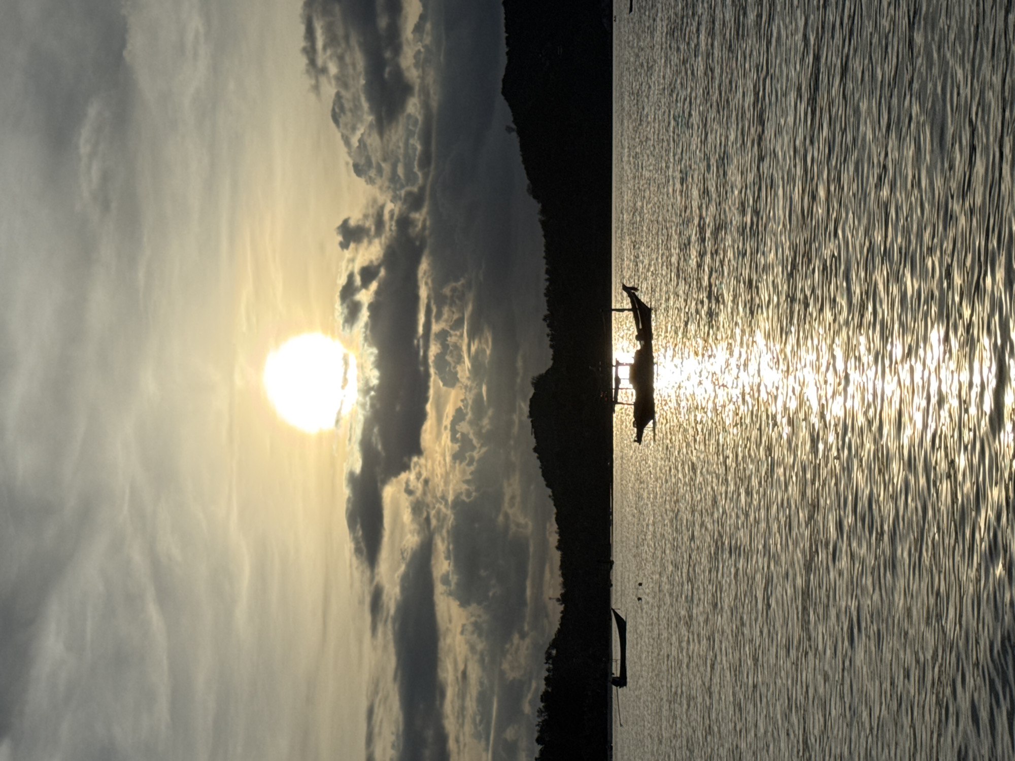 The image depicts a tranquil sunset scene over the waters of Koh Phangan, Thailand. A bright, hazy sun is partially obscured by textured clouds in the upper portion of the frame. The sun's light casts a shimmering golden reflection across the choppy surface of the sea. Silhouetted against the horizon are the dark outlines of a lush, tree-covered island. Several small boats are visible on the water. In the foreground, a long, narrow boat with a canopy is anchored. Further in the distance, another boat is moored, and a few small, dark figures, possibly swimmers or floating objects, are present. The overall atmosphere is serene and natural, characteristic of a coastal sunset in Southeast Asia.