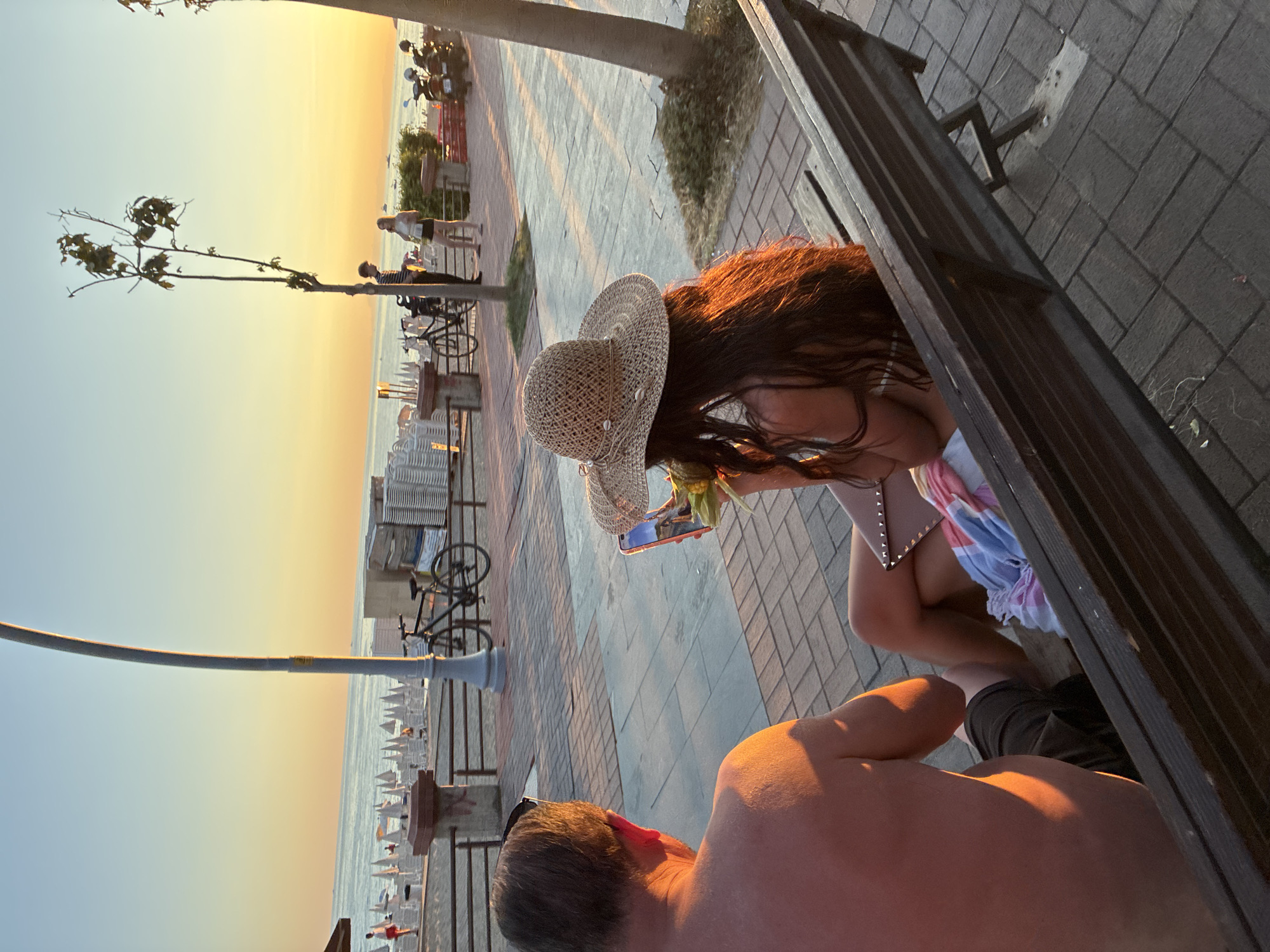 Two individuals are seated on a wooden bench on a paved walkway adjacent to a beach. The person on the left, an adult male with light skin and short hair, faces away from the camera. He is shirtless and wears dark shorts. The person on the right, an adult female with long dark hair, faces left, looking at a smartphone. She wears a wide-brimmed straw hat and a bikini top. A colorful towel is draped over her lap.
In the background, the beach is visible with rows of lounge chairs and umbrellas. The sea stretches to the horizon, under a sky with a gradient of orange and blue hues, indicating sunset. Various objects are scattered along the walkway and the beach, including bicycles, scooters, and stacks of chairs. The location appears to be the Alanya waterfront in Turkey, suggested by the distinctive architecture of the lampposts and the general ambiance of a Mediterranean beach resort at dusk.