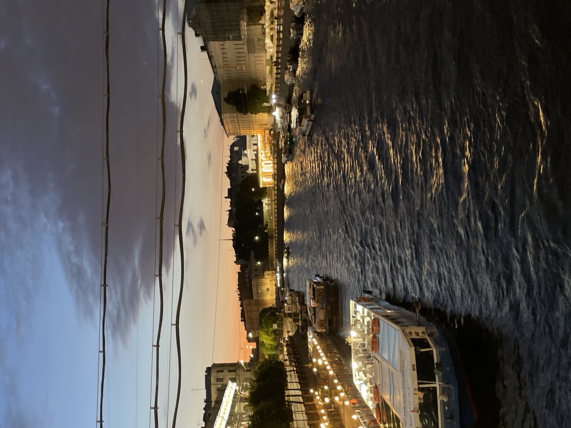 The image shows a scenic view of a canal in Saint Petersburg, Russia at dusk. The sky transitions from a pale blue at the top to hues of orange and pink near the horizon, indicating either sunrise or sunset. Several buildings with illuminated windows line the canal, with trees and streetlights visible along the embankment. A prominent feature is the presence of numerous boats navigating the water. In the foreground, a large passenger boat with multiple decks and rows of seats is docked or moving slowly. Other boats, including smaller tour boats and what appear to be barges, are also visible further along the canal. The water reflects the lights from the buildings and boats, creating a shimmering effect. Overhead, power lines or cables stretch across the frame, obscuring a small portion of the sky. The overall atmosphere is serene and picturesque, capturing the charm of Saint Petersburg's waterways during the evening.