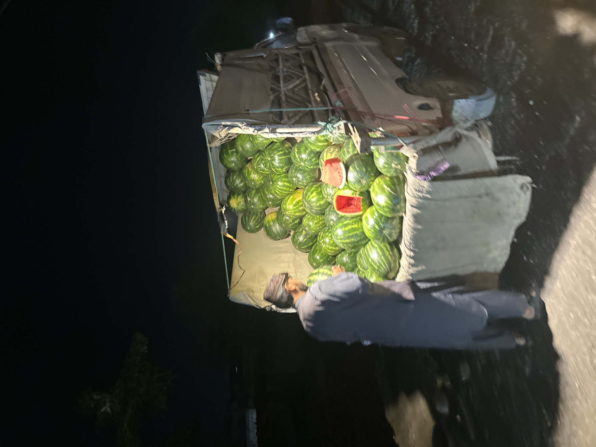 The image captures a nighttime scene, likely in Quetta City Tehsil, Pakistan, focused on a roadside vendor and his produce. A man, dressed in a dark shirt and pants with a beard and a cap or head covering, stands beside the rear of a parked pickup truck or small lorry. The truck's bed is heavily loaded with numerous green-striped watermelons, some of which are cut open to reveal their red flesh, suggesting they are being displayed or checked for quality. The vehicle has a makeshift cover or canopy over its cargo area. The background is dark, with faint outlines of trees on the left, indicating an outdoor setting. The scene is illuminated by artificial light sources, casting shadows and highlighting the watermelons and the man. The activity appears to be the sale, inspection, or preparation of watermelons from the truck. No discernible text is visible in the image.