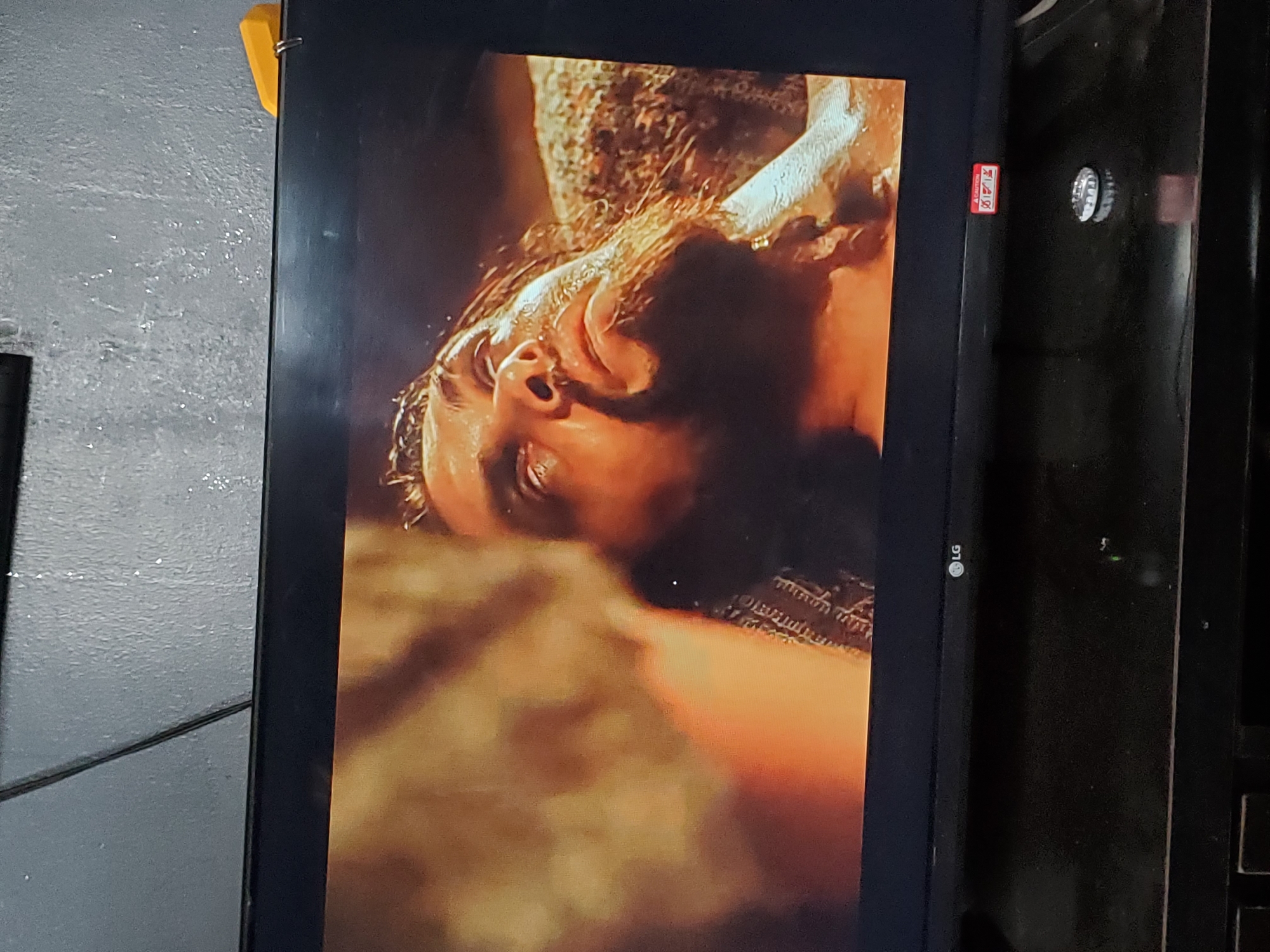 The image shows a close-up of a television screen displaying a scene from a movie or show. The central subject is a man with a beard, lying down and looking upwards, with his eyes wide open. His skin appears wet, and there are details of his hair and the texture of his clothing visible. The background of the image on the screen is blurry and appears to be in warm, earthy tones. The television itself is black with an "LG" logo visible on the bottom right. The television is placed on a dark, reflective surface, possibly a piece of furniture. On the surface in front of the TV, there is a metal bottle cap and a red and white sticker. The wall behind the television is textured and appears to be gray. The image does not provide any specific information about the location in Winters, United States of America, or any context for the scene being viewed.