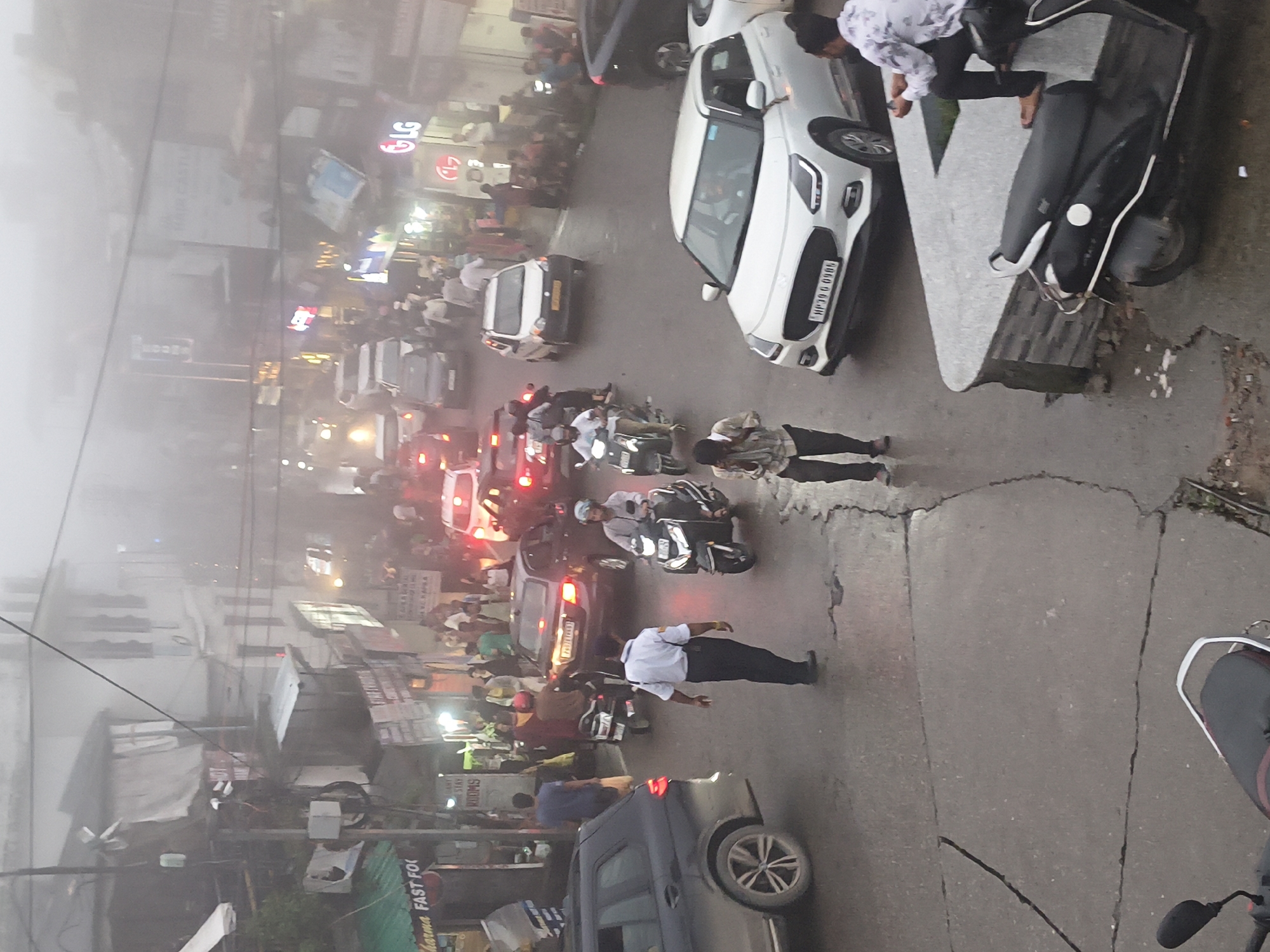 This is an eye-level, high-angle, full shot of a foggy street scene in Dharamshala, India, possibly during monsoon season given the wet road surface and haze. A variety of vehicles including cars and scooters are present, indicating moderate traffic. Several individuals are observed walking on the street and pavements, some are wearing casual clothing, while one individual in a light blue shirt and dark pants appears to be directing traffic. Commercial establishments with visible signage are lined up along the street, contributing to a bustling urban environment. The overall atmosphere is hazy, suggesting poor visibility, which is characteristic of atmospheric conditions in the region.