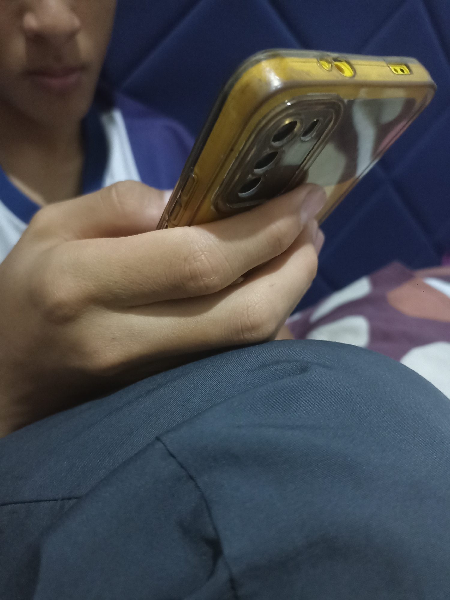 A close-up shot shows a person's hands holding a smartphone in Mojopanggung, Indonesia. The person is wearing a blue and white shirt. The smartphone has a yellow case with a camera module on the back. The background is blurred, but appears to be an indoor setting with a blue textured surface and a patterned pillow. The person's face is partially visible at the top of the frame, and their hands are positioned to operate the phone. The overall impression is one of casual use of a mobile device.