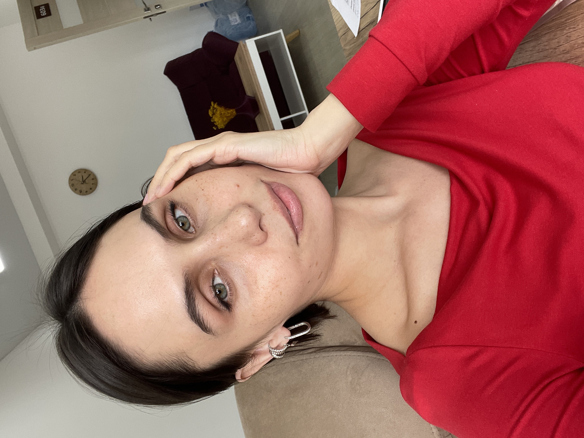 The image captures a young woman taking a selfie indoors, likely in Izhevsk, Russia. She is shown lying down, looking directly at the camera with a gentle smile, her left hand resting lightly on her forehead. She has dark hair, prominent green eyes, and freckles visible on her forehead, nose, and cheeks. She is wearing a long-sleeved, form-fitting red top. Her left ear features two silver-colored earrings: a multi-band ear cuff and a carabiner-style or heart-shaped hoop.
The background indicates an indoor setting, possibly a home or office. A light-colored wall with a small, round wooden clock is visible. Part of a light wooden door frame is in the upper background, displaying the number "6036." Other elements include a dark red couch or chair, a white shelving unit with some items, and a large bottle of water on a distant surface. The lighting appears bright, suggesting it is daytime. The photograph is taken from an angled, rotated perspective, contributing to a relaxed and candid feel.