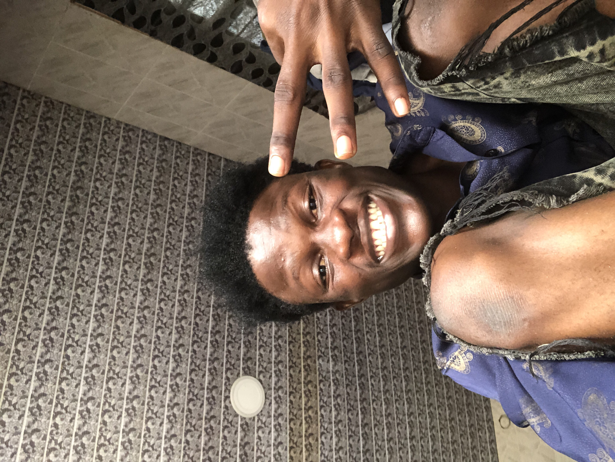 A young man with dark skin and short, dark, curly hair smiles broadly at the camera, holding up two fingers in a peace sign with his right hand. He is wearing a dark blue shirt with a gold paisley pattern and appears to be in a room with textured walls. One knee, visible in the foreground, is also showing the texture of his skin. The image seems to have been taken in Ajah, Nigeria.