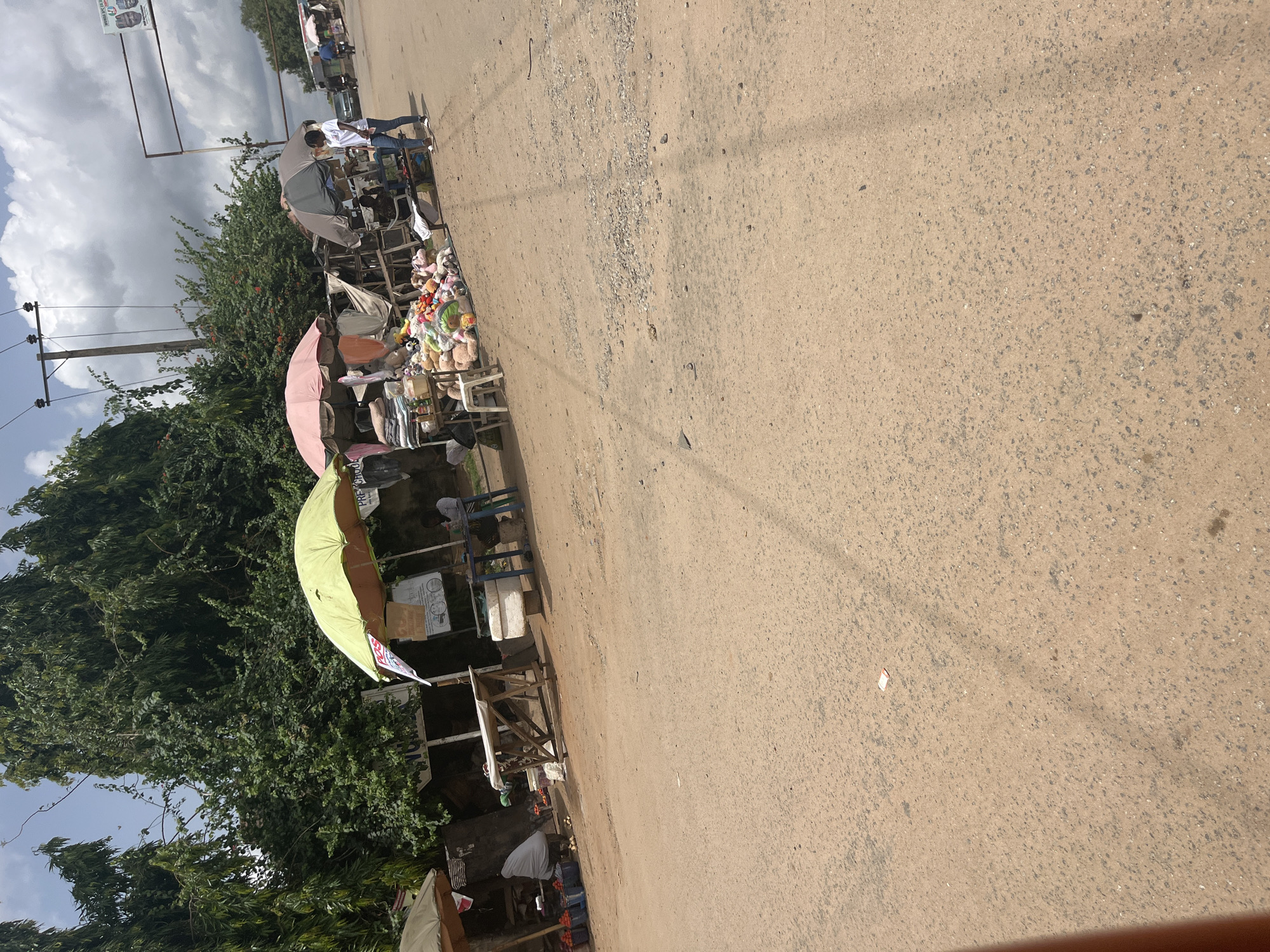 A street in Abuja, Nigeria, features vendors selling various goods. The road is unpaved and dusty. Along the side of the road, several stalls are set up, shaded by umbrellas of different colors (green, pink, gray). People are visible near the stalls, potentially interacting with the vendors. Goods are displayed on tables, including what appear to be stuffed animals and other items. Trees and a utility pole with wires are also visible. The sky is cloudy.
