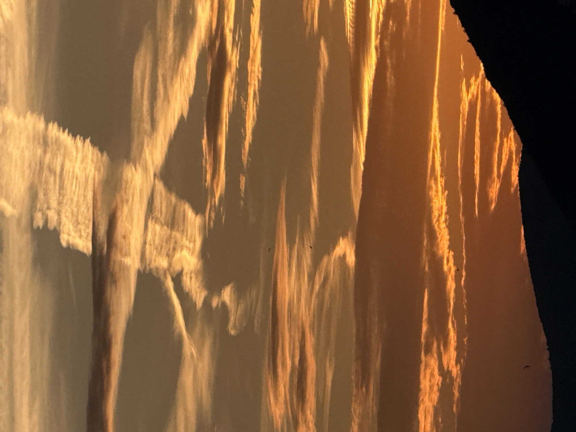 A silhouetted mountain range borders the bottom of the frame, against a sky ablaze with the warm hues of sunset. Wispy clouds, tinged with orange and gold, stretch across the upper portion of the image, forming intricate patterns. A contrail, a stark white line against the warmer tones, cuts a diagonal path through the sky. Two birds are visible in flight, small dark specks against the vibrant backdrop, adding a sense of scale and life to the expansive sky. The overall scene evokes a sense of natural beauty and tranquility at the end of the day. This image appears to have been captured in the vicinity of Pržno, Montenegro, given the characteristic silhouette of the coastal mountains.