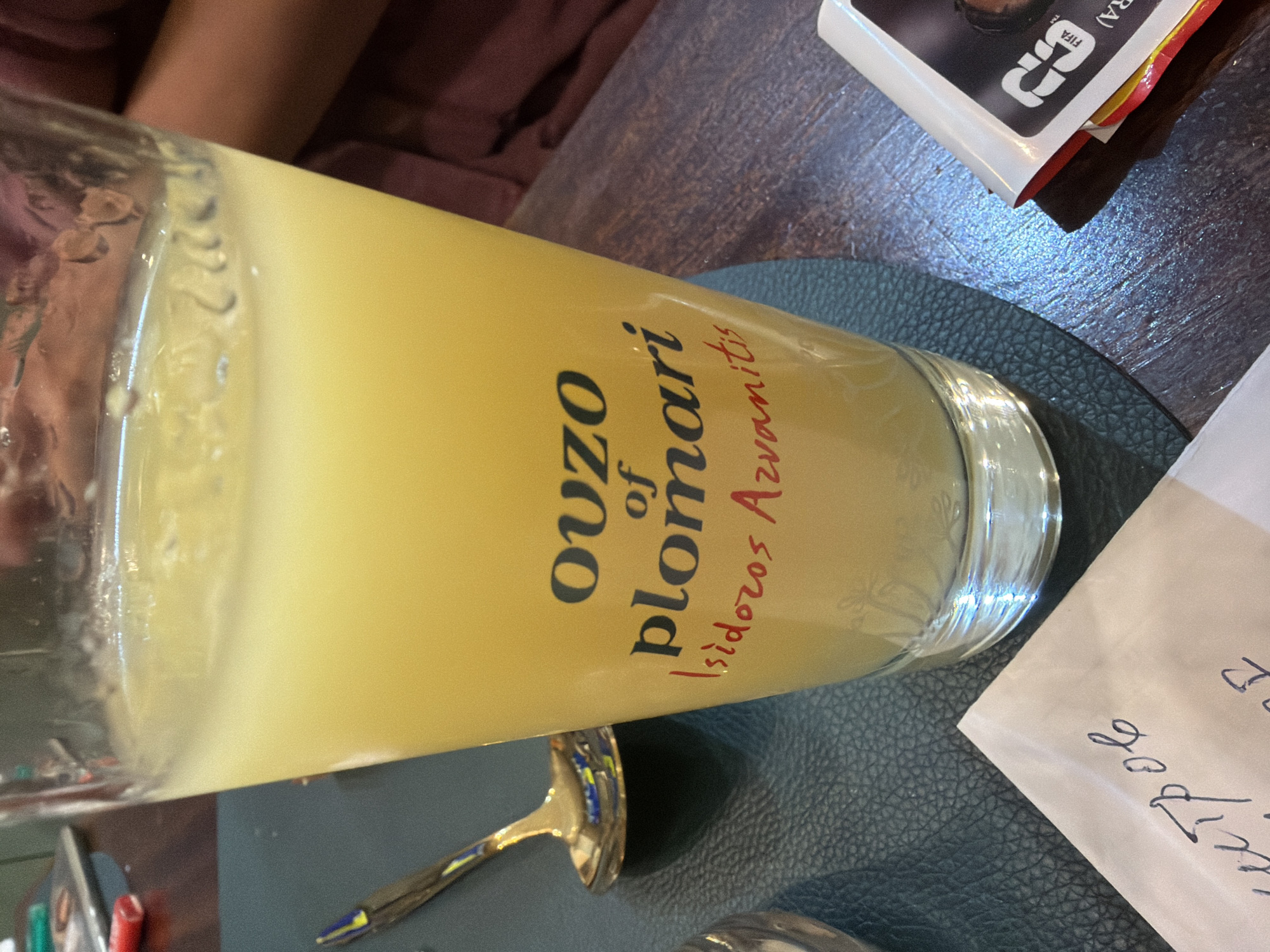 A close-up shot shows a glass of pale yellow liquid, likely an alcoholic beverage, with a label that reads "OUZO of plomari Isidoro Avianitis." The glass is on a dark, textured surface. To the left of the glass is a metallic spoon. In the foreground, a corner of a paper napkin with handwritten text is visible. To the right of the glass, a small stack of items, including a white card with "FIFA" branding and a partial image of a person, are present. The background is blurred, showing a hint of someone's arm and shoulder wearing dark clothing. The lighting appears to be ambient, with some reflections on the glass and table surface. The location is not specified.