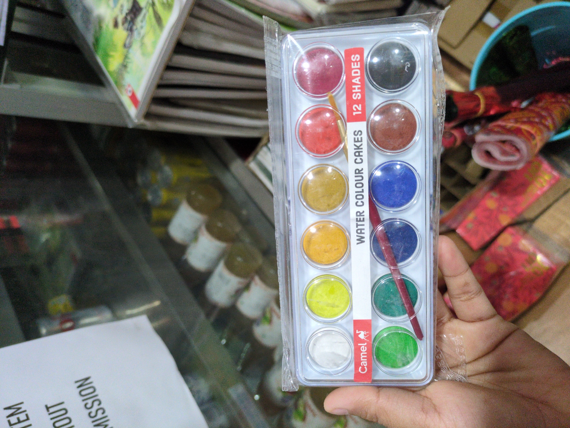 The image shows a close-up of a hand holding a transparent plastic package containing a 12-shade watercolor paint set by Camel. The paint set is positioned in the foreground, with the individual paint cakes visible through the plastic. The brand name "Camel" and the text "WATER COLOUR CAKES 12 SHADES" are clearly legible. A thin paintbrush with a reddish-brown handle rests across some of the paint cakes. In the background, shelves are visible, displaying various items that appear to be in a shop or market setting, consistent with a location in Bhagsu, India, likely a small store selling art supplies or general goods. The overall lighting suggests an indoor environment.
