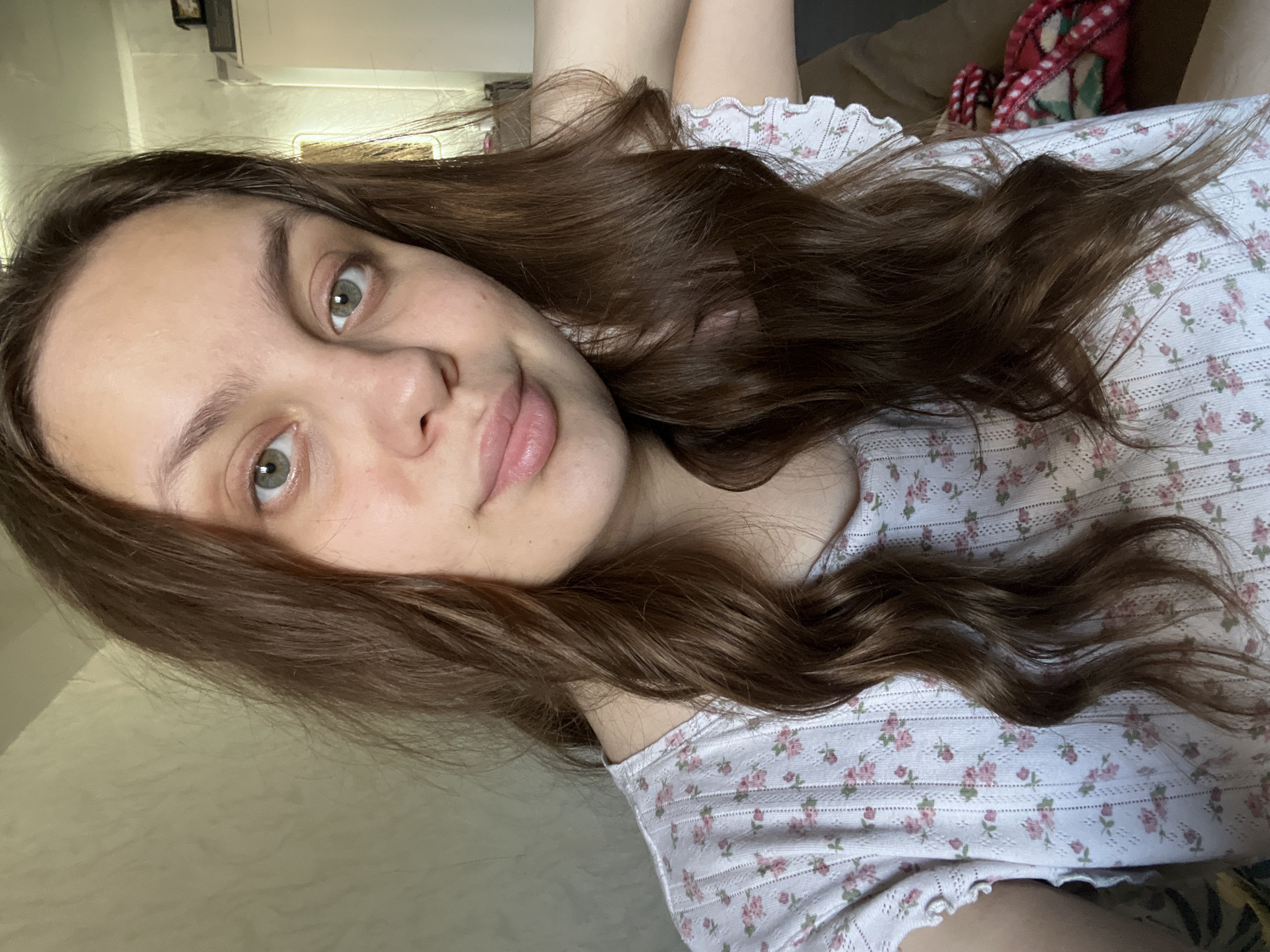 A young woman with long, wavy brown hair is reclining and taking a selfie. She has fair skin, green eyes, and is wearing a light-colored floral print top. The background is a slightly blurred interior, likely a room in a residence. A blanket or covering with a red and white pattern is partially visible behind her. The location is not identifiable as Chaikovsky, Russia.