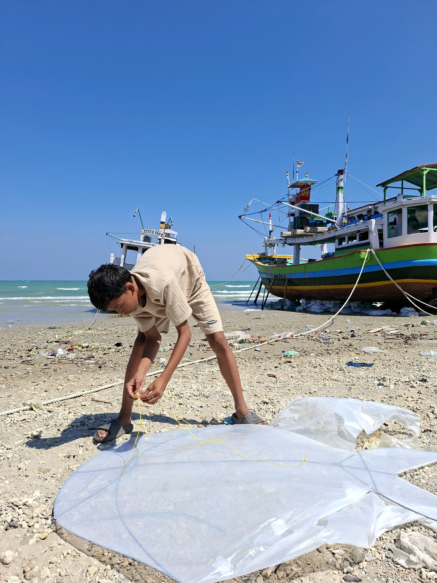A young boy with dark hair, wearing a tan t-shirt and shorts, is on a beach in Camplong, Indonesia. He is bent over, focused on attaching yellow strings to a large, white kite laid out on the sand. The kite appears to be made of thin plastic material and has a star-like or fish-like shape with visible internal struts. He is wearing dark grey sandals on his feet.
The beach is covered in sand, small stones, and debris, including plastic bottles and wrappers, suggesting it is not entirely clean. In the background, two boats are visible. A larger, colorful fishing boat with green, blue, yellow, and brown hull is propped up on stilts on the sand. Behind it, a smaller white boat with a red and white flag is partially visible, also on the shore. The sea is a clear turquoise color with gentle waves lapping at the shore, and the sky above is a bright, cloudless blue. The sun is shining, casting a shadow of the boy on the sand.