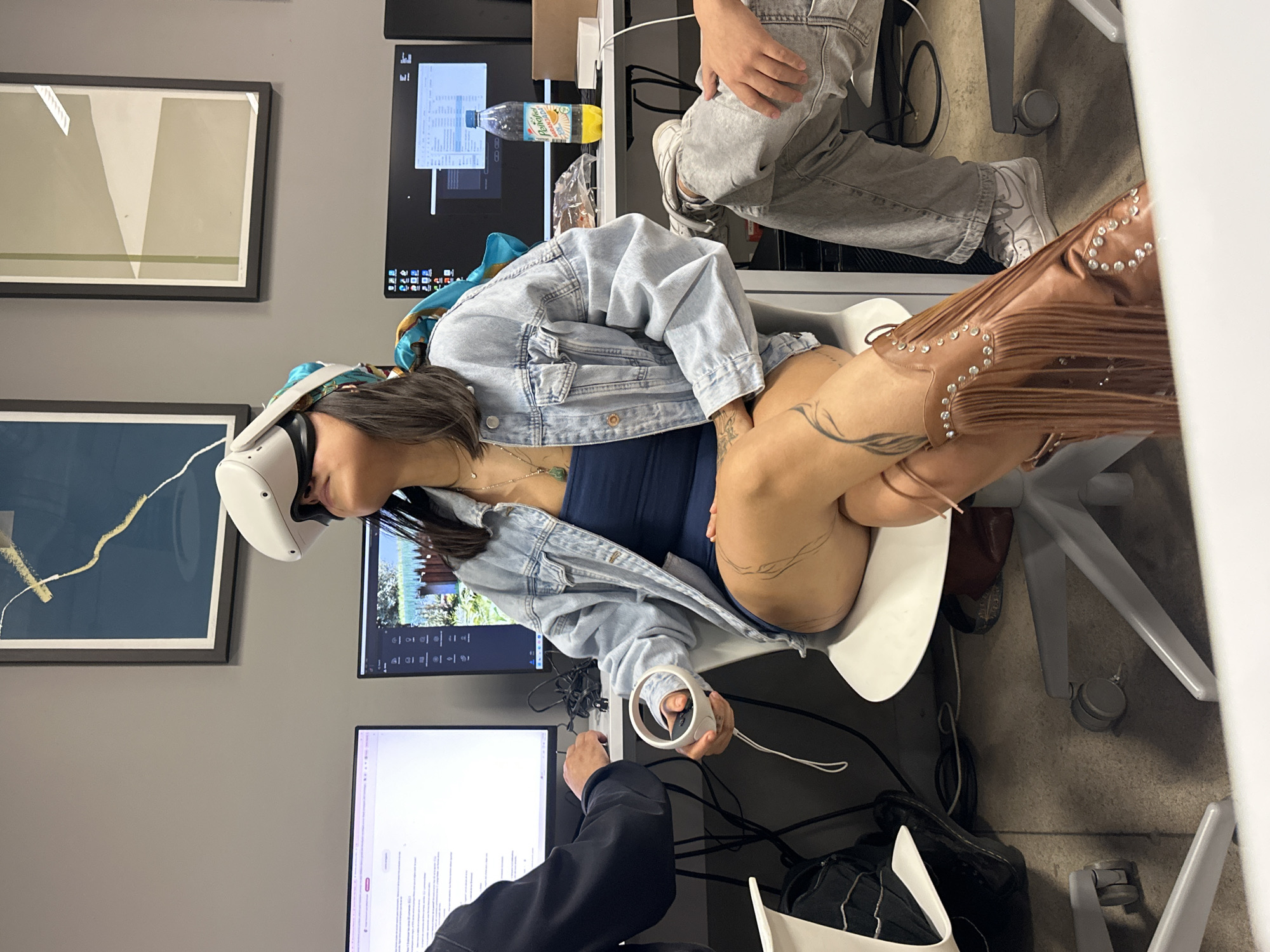 The image shows a woman wearing a VR headset and holding a controller in an office setting. She is seated in a white chair and wearing a blue top, denim jacket, and fringed brown boots. Tattoos are visible on her leg. There are at least two computer monitors on the desk in front of her. A person is partially visible to the right of the woman in light grey jeans and white sneakers. Another person in a black jacket is visible to the left. Two framed artworks hang on the grey wall behind the woman. A bottle of Jumex juice sits on the desk. There is no information to prove the location is in Mexico City, Mexico.