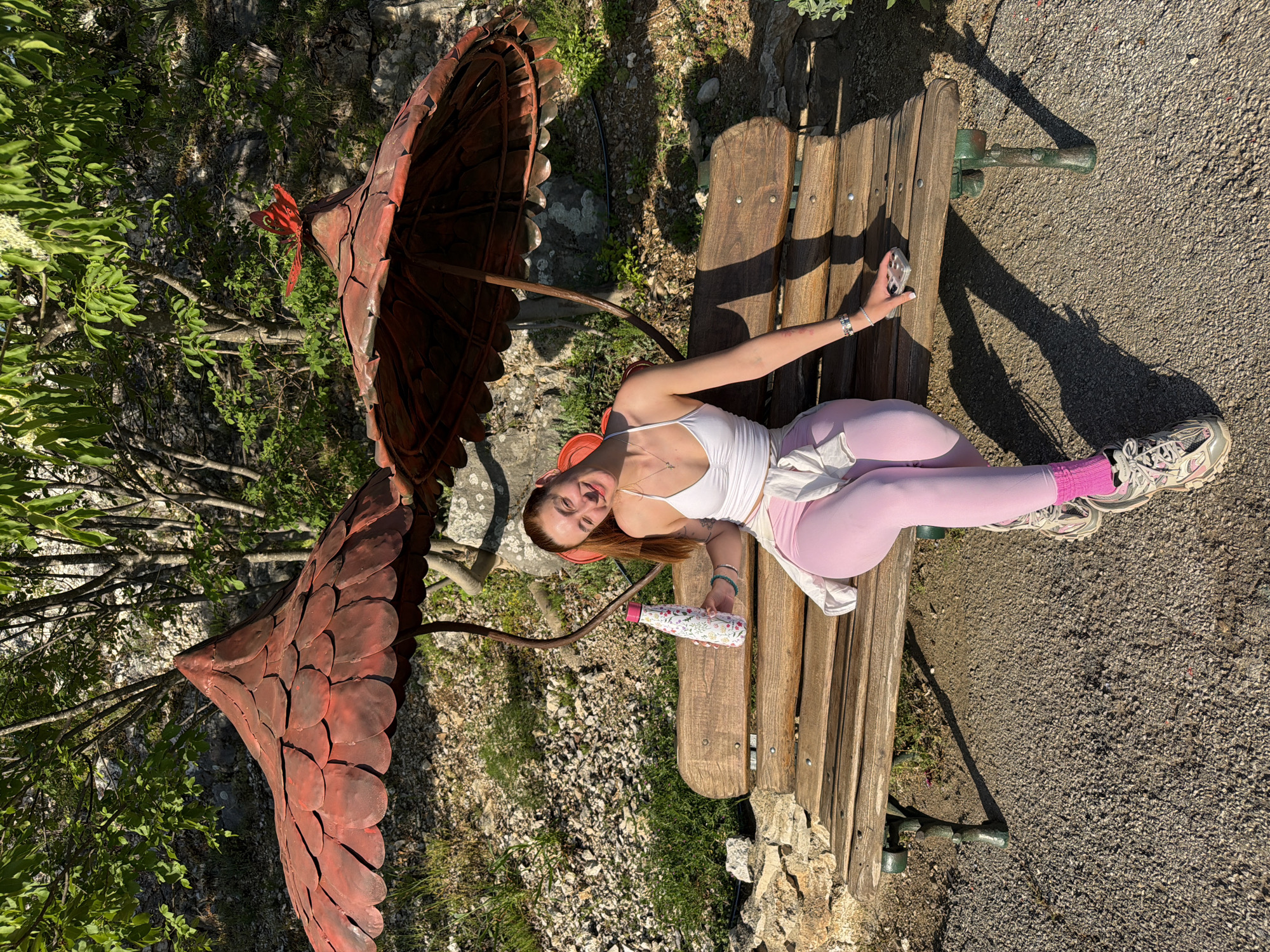 A full shot depicts a fair-skinned female lying on a wooden park bench, wearing a white tank top and light purple leggings. She's in a relaxed position with her arms outstretched; in one hand, she holds a patterned water bottle, and in the other, a cell phone. She sports pink socks and athletic shoes.
To the left of the bench is a large, ornate metal sculpture resembling a mushroom. The background includes a mix of vegetation and rocky terrain. The location is identified as Lukovit, Bulgaria. The image has a warm, sunny atmosphere.