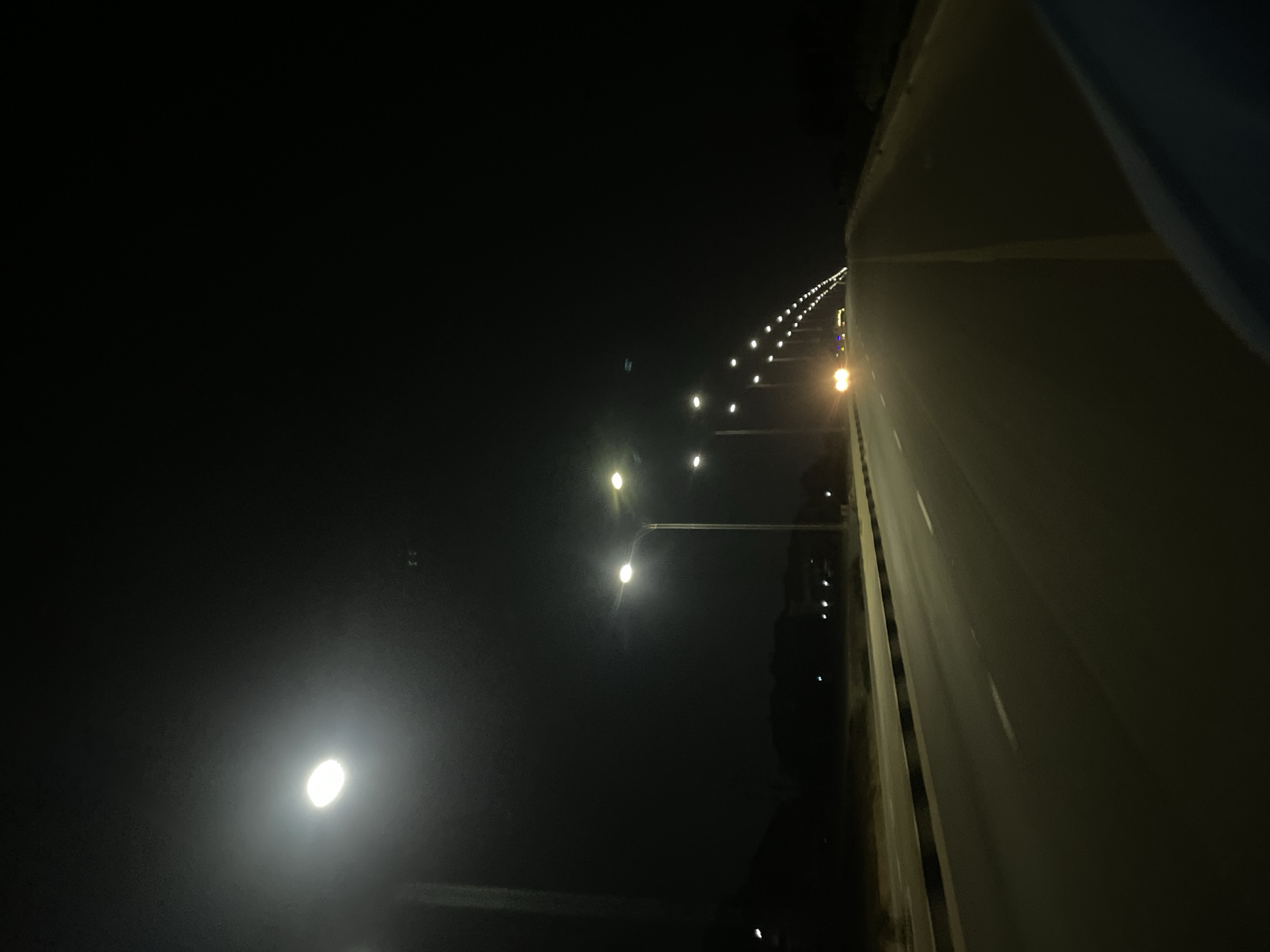 The image shows a night scene along a road in Dam Camp, Nigeria. Streetlights illuminate the road, which stretches into the distance. The area beyond the road appears dark. A vehicle with its headlights on is visible further down the road. The road surface is visible, and there appears to be a barrier or shoulder to the right. No people are visible in the immediate foreground.