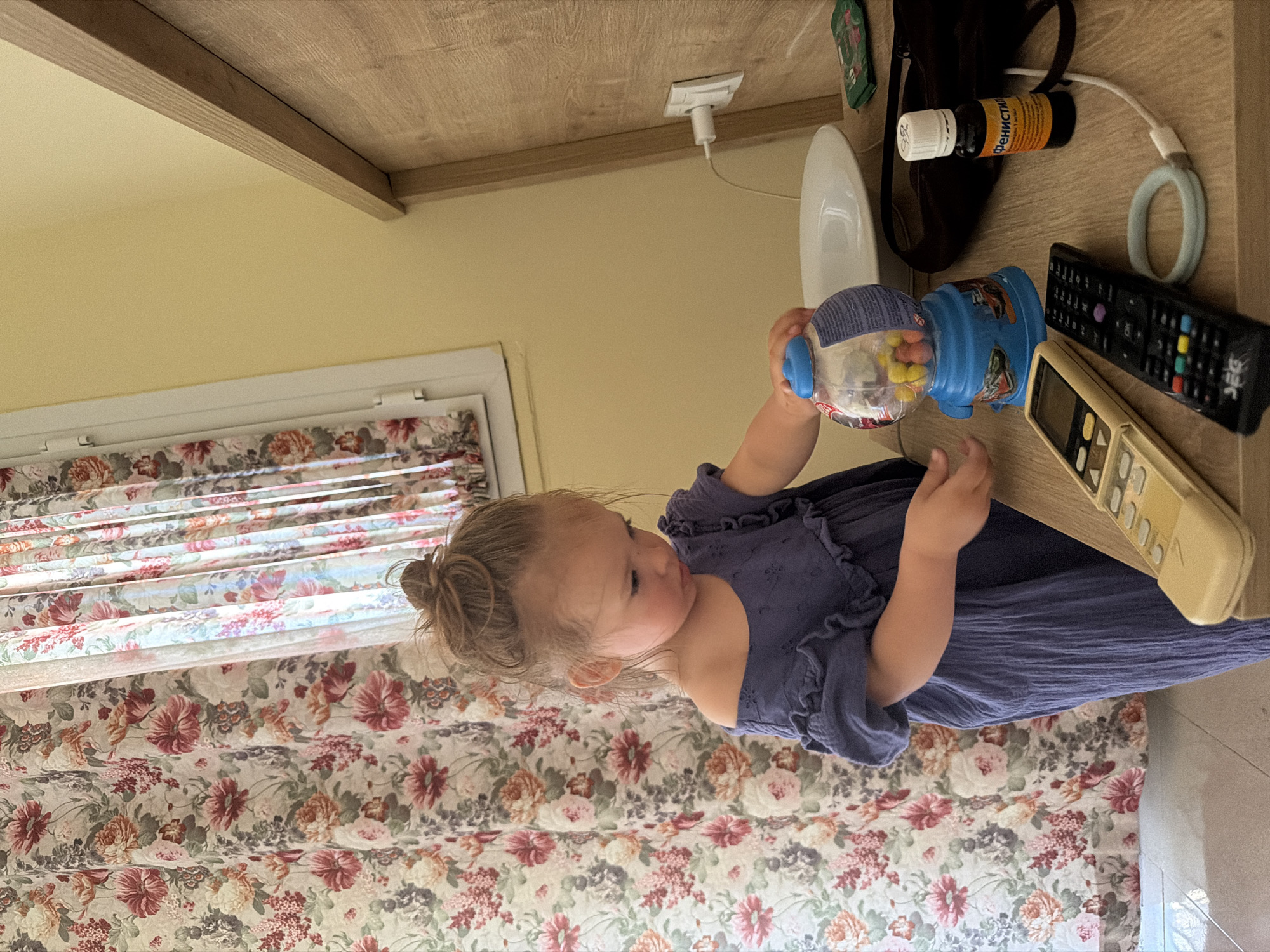 A young girl with her hair in a bun is standing next to a floral curtain and a wooden headboard. She is wearing a blue dress and is playing with a blue gumball machine. In front of her, on a wooden table, are two remote controls, a small bottle of medication, and a white ring. A white electrical outlet with a white cord is visible on the wall behind the table. The room appears to be a bedroom in a hotel located in Fethiye, Turkey.