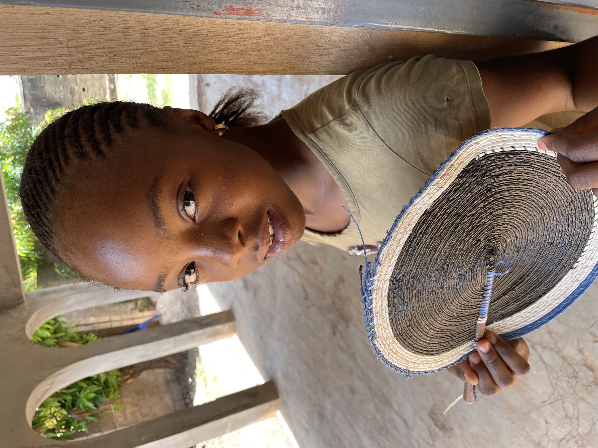 A young woman with braided hair and a gold earring holds a woven circular object with a handle. The object features a pattern of alternating dark and light bands, with a blue border. The woman is wearing a light green t-shirt and is positioned under a wooden structure. The background includes concrete pillars and greenery, suggesting an outdoor or semi-outdoor space. The location is likely within the city of Jos, Nigeria.